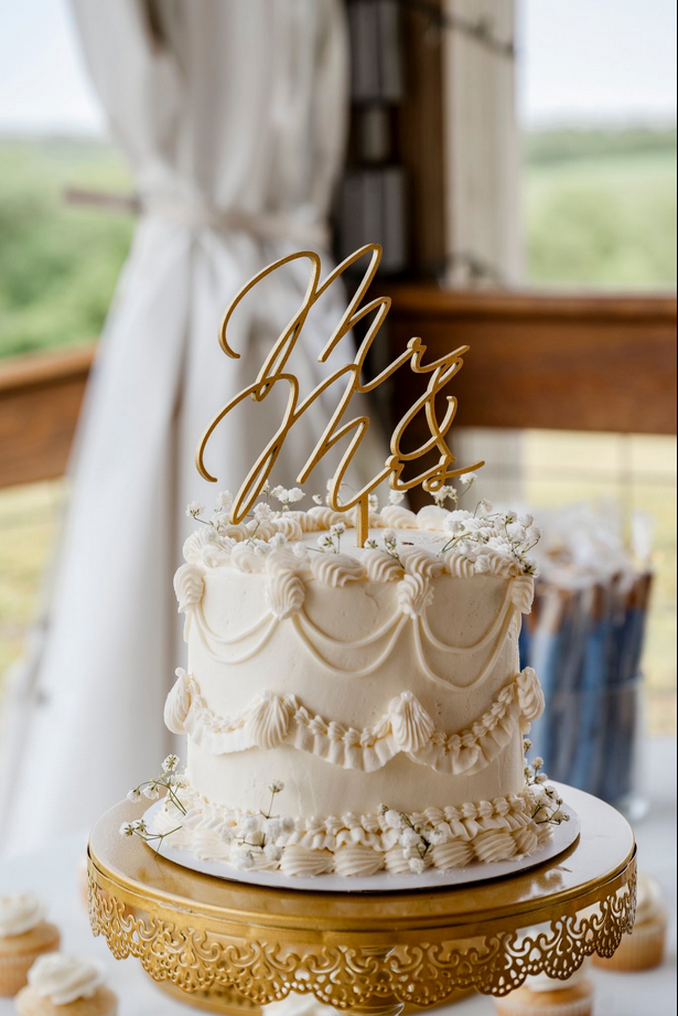 Cream-colored wedding cake with decorative swirls and floral accents, topped with a gold.  Dream Day Event Rentals, Vintage and Modern Decor and Floral Rentals, Freeport Illnois.