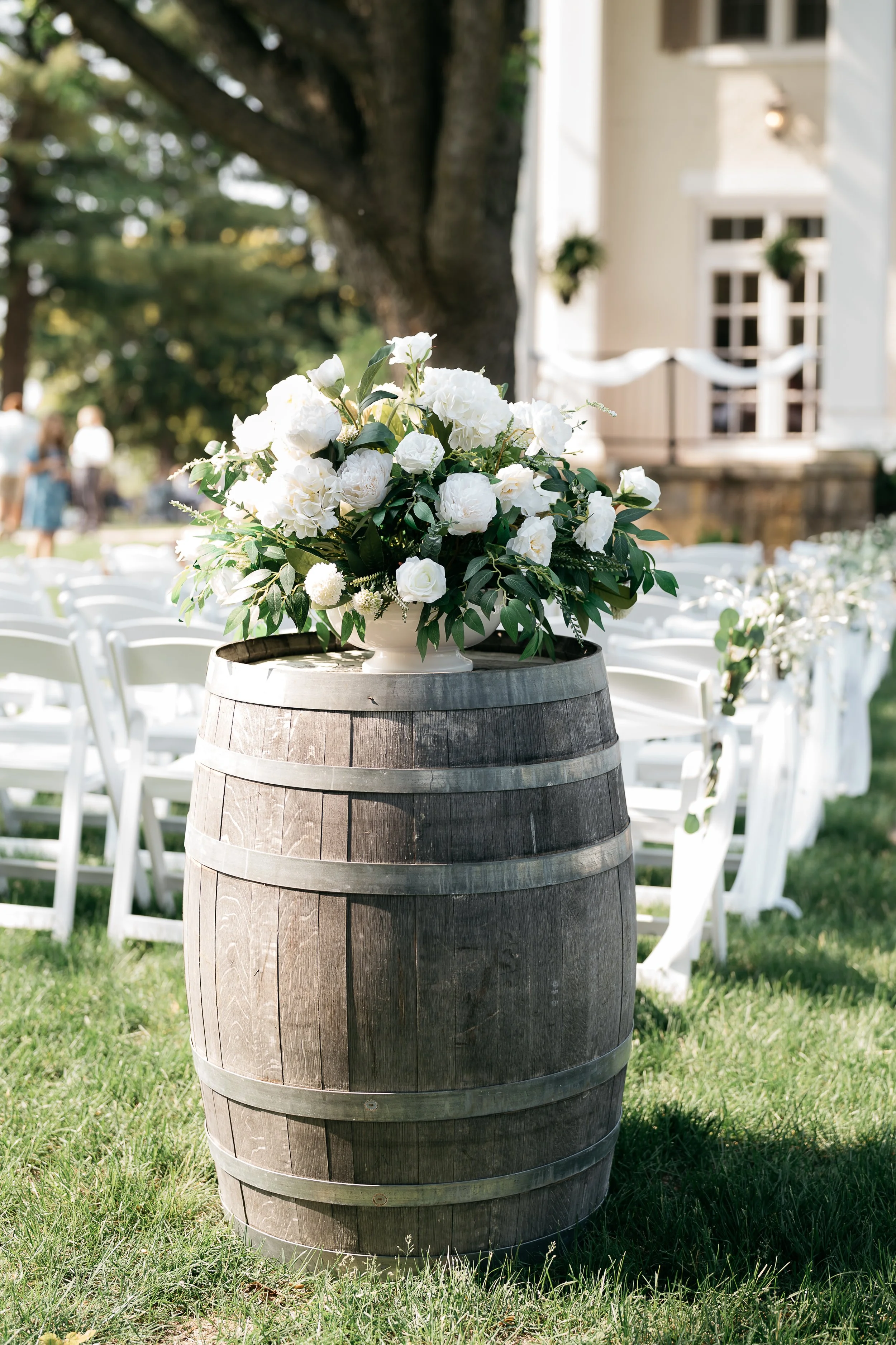 A large wooden barrel with a white flower arrangement on top, set outdoors near white garden chairs. Dream Day Event Rentals, Vintage and Modern Decor and Floral Rentals, Freeport Illnois and Southern Wisconsin