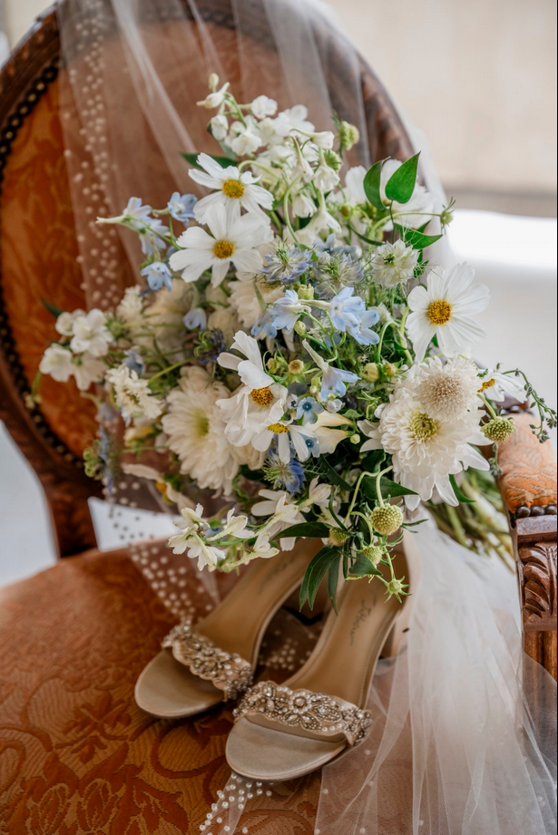 Wedding bouquet of white daisies, blue delphiniums, white dahlias, and greenery resting on a pair of beige high-heeled shoes with rhinestone embellishments, placed on an ornate wooden chair with a sheer veil draped over it.