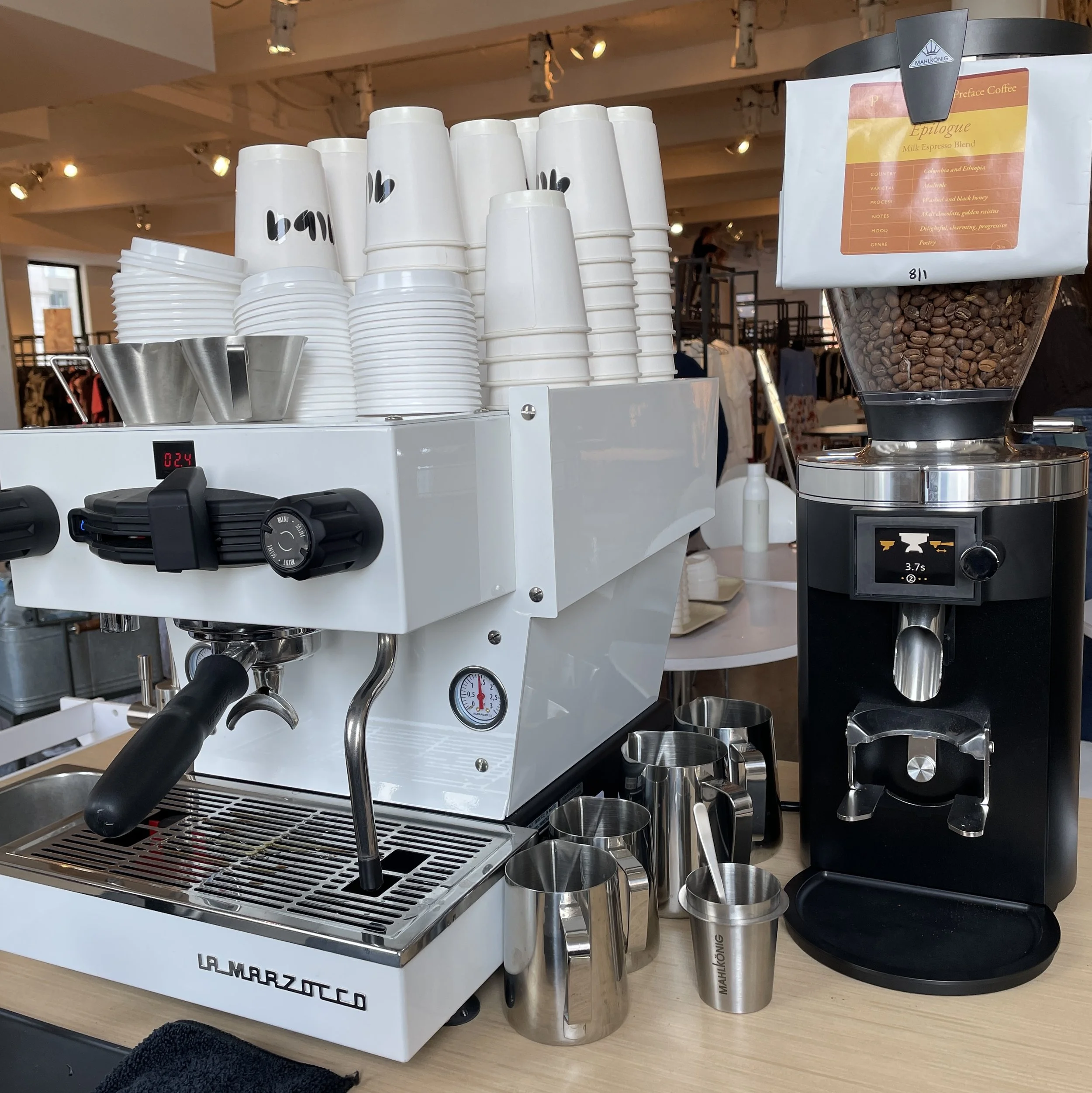 A white La Marzocco espresso machine with cups, bowls, and milk frothing pitcher on a countertop. To the right, a grinder filled with coffee beans and a bag of preground coffee are visible.