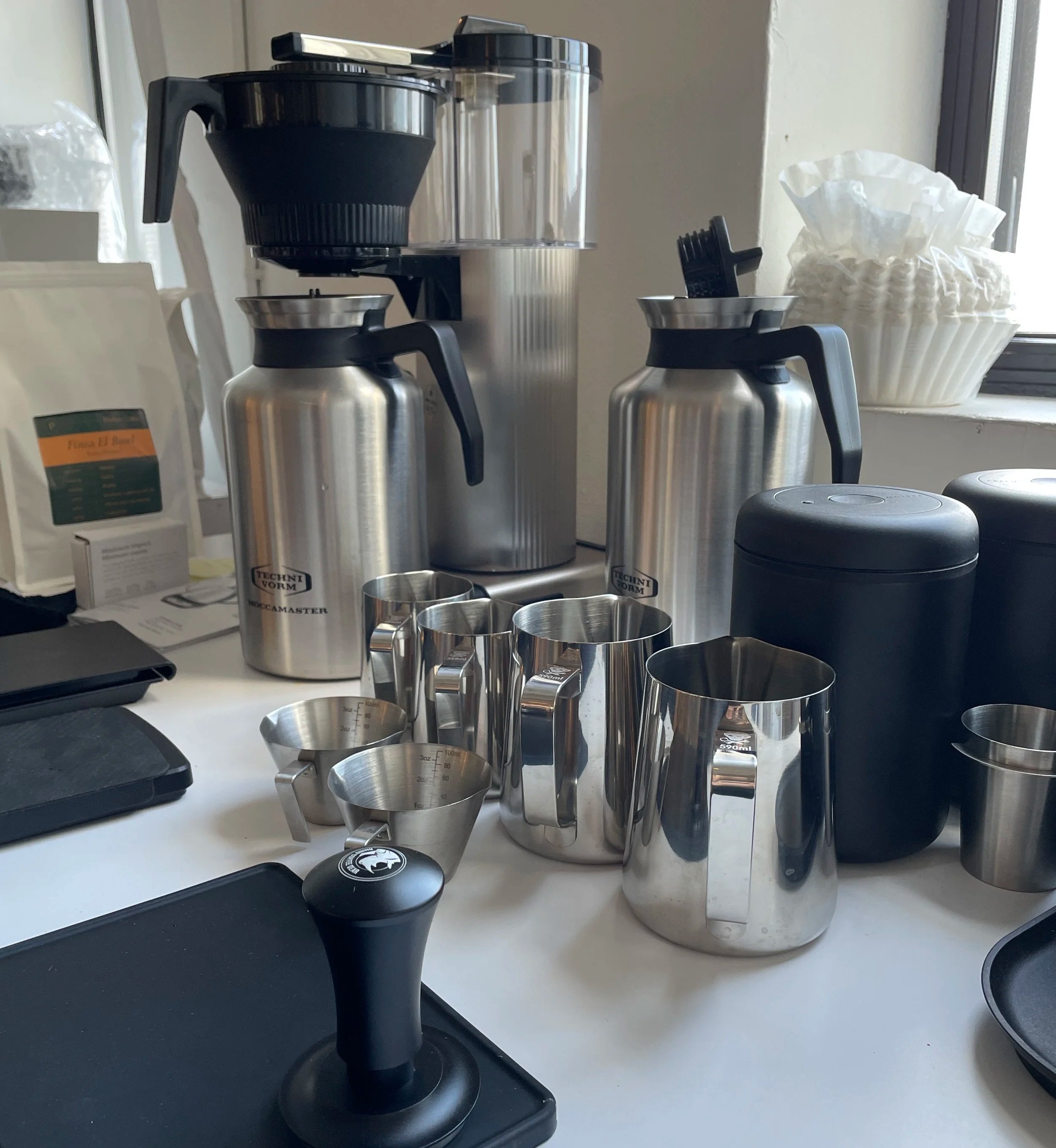 A coffee station with a coffee maker, stainless steel pitchers, cups, and coffee accessories on a white countertop near a window.