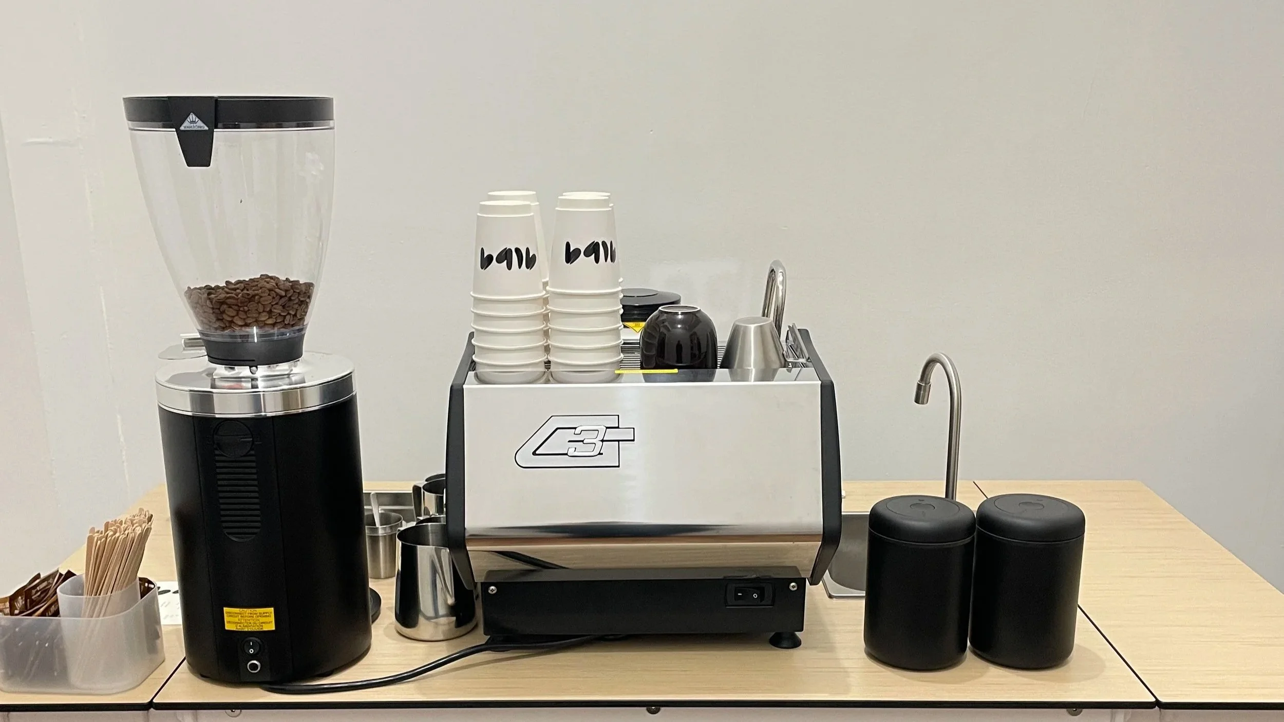 Coffee station with a coffee grinder, cups, a coffee machine, and containers for coffee supplies on a wooden table.