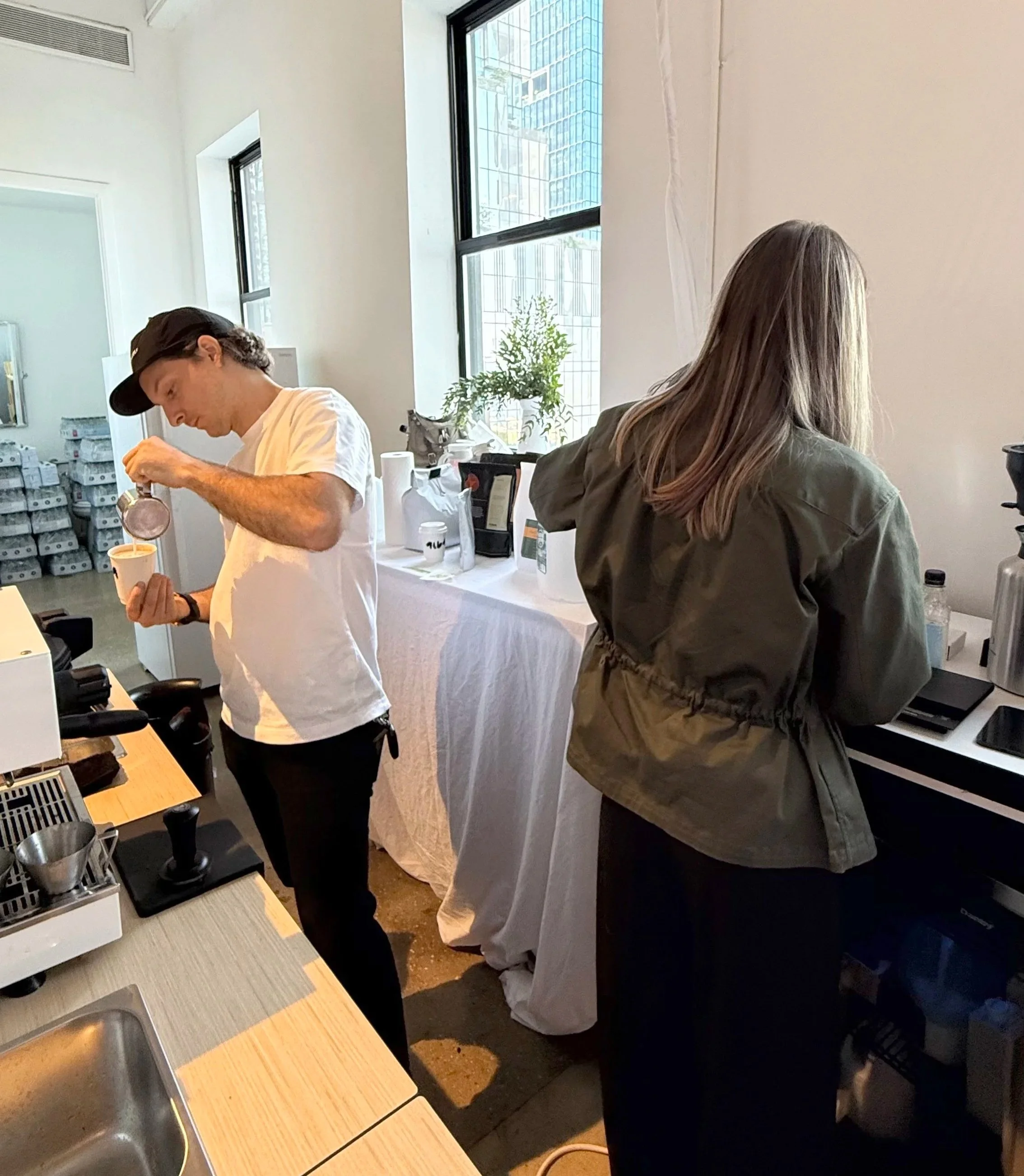 A first barista is pouring coffee into a cup while a second stands at a counter with a coffee machine in a bright cafe with large windows.