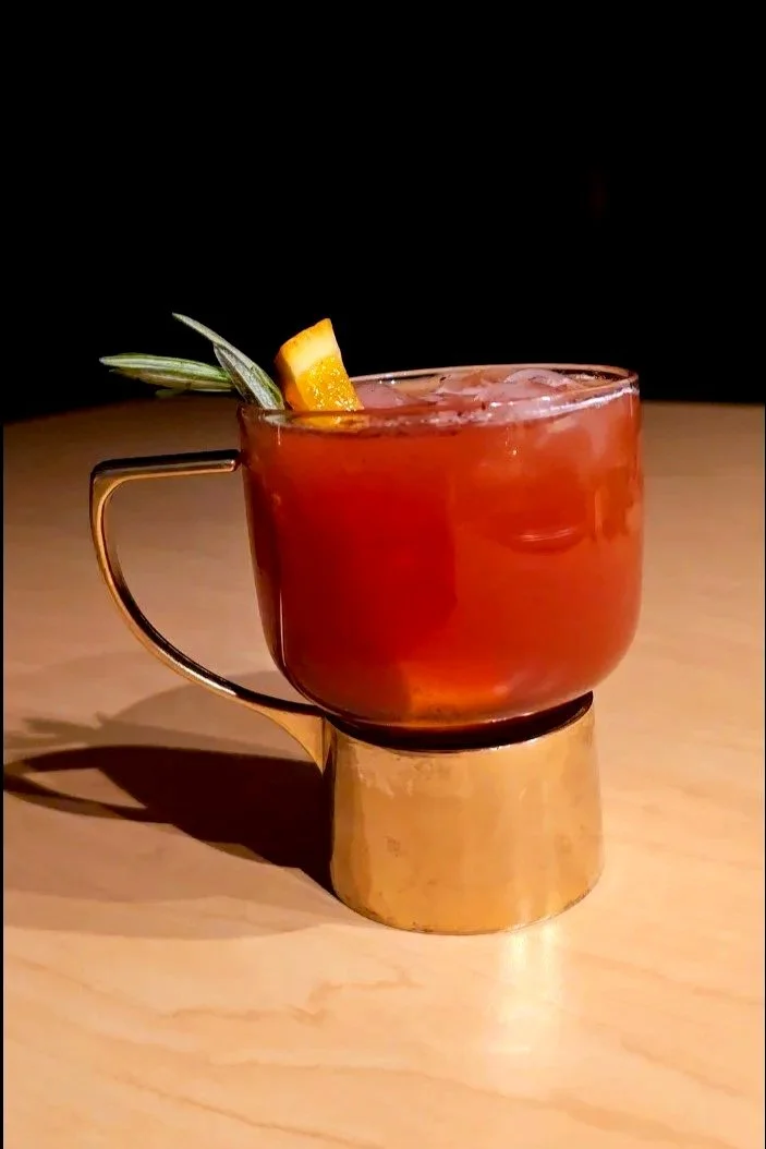 A red-colored cocktail in a transparent glass cup with a gold handle, garnished with a pineapple leaf and an orange slice, placed on a round gold stand on a wooden surface.