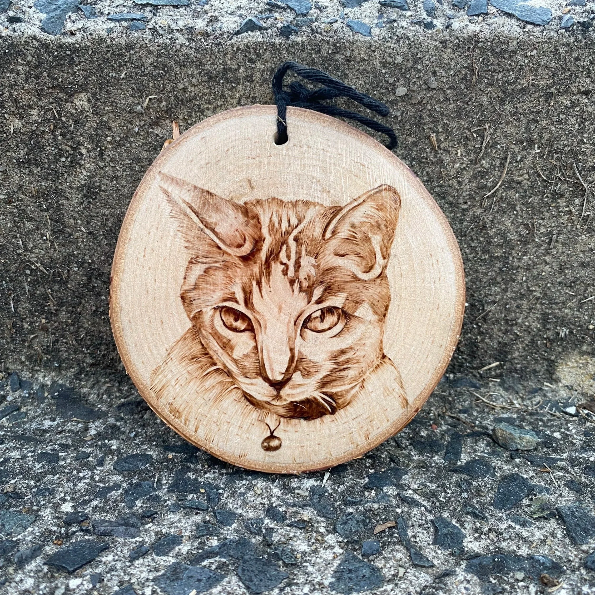 Wooden ornament with a detailed engraving of a cat's face, hanging on a black string, placed on gravel and concrete ground.