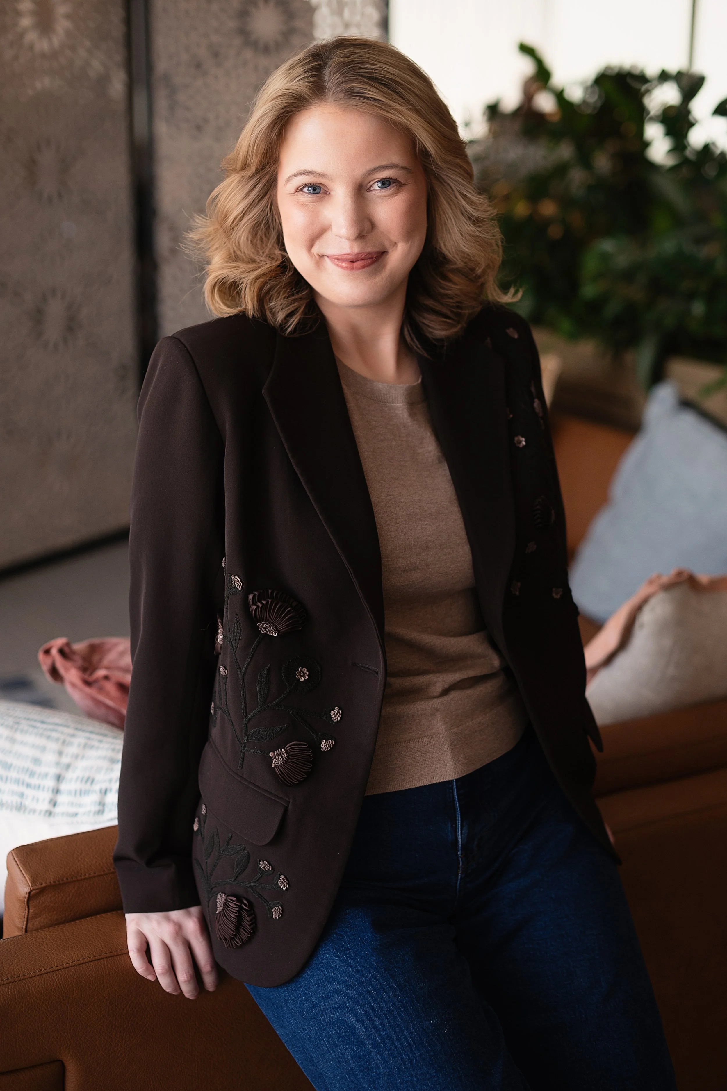 A woman with blonde hair and blue eyes, smiling, wearing a black embroidered blazer, beige shirt, and blue jeans, sitting in a cozy, well-lit room.