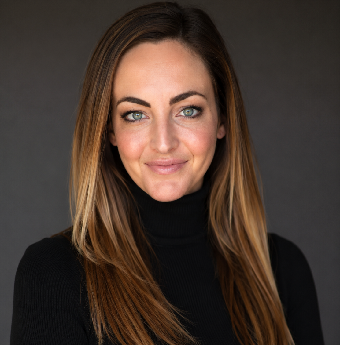 Close-up portrait of a woman with long brown hair, wearing a black turtleneck, smiling gently against a dark background.