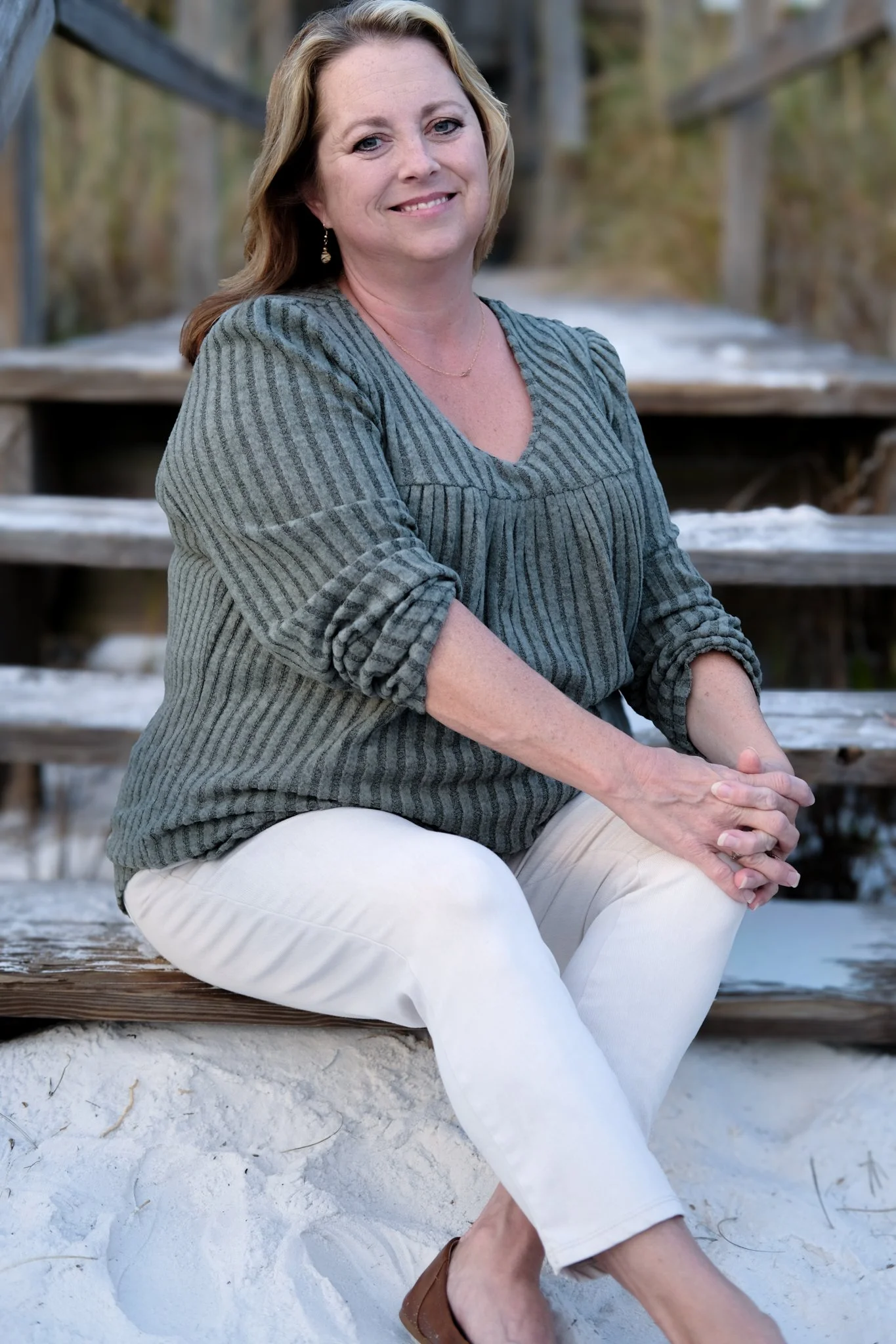 A woman sitting on wooden steps outdoors, smiling with hands clasped on her knee, wearing a gray striped blouse and white pants.