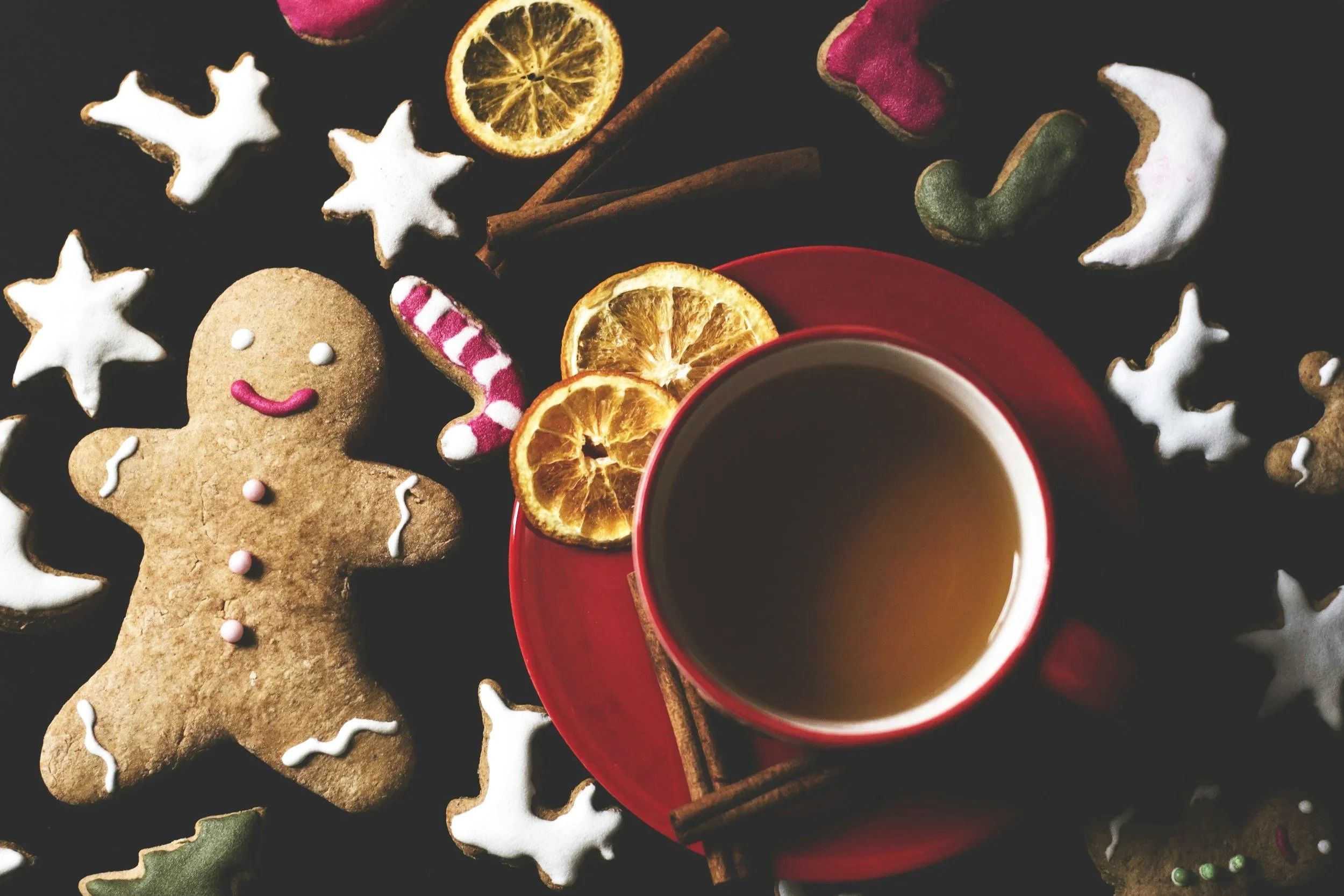 A red cup filled with a hot beverage, placed on a red saucer. Surrounding the cup are dried orange slices, cinnamon sticks, and Christmas-themed cookies decorated with white icing, including a gingerbread man with pink buttons and a smile. There are also other cookies shaped like stars and Christmas trees, some decorated with red or green icing and sprinkles.