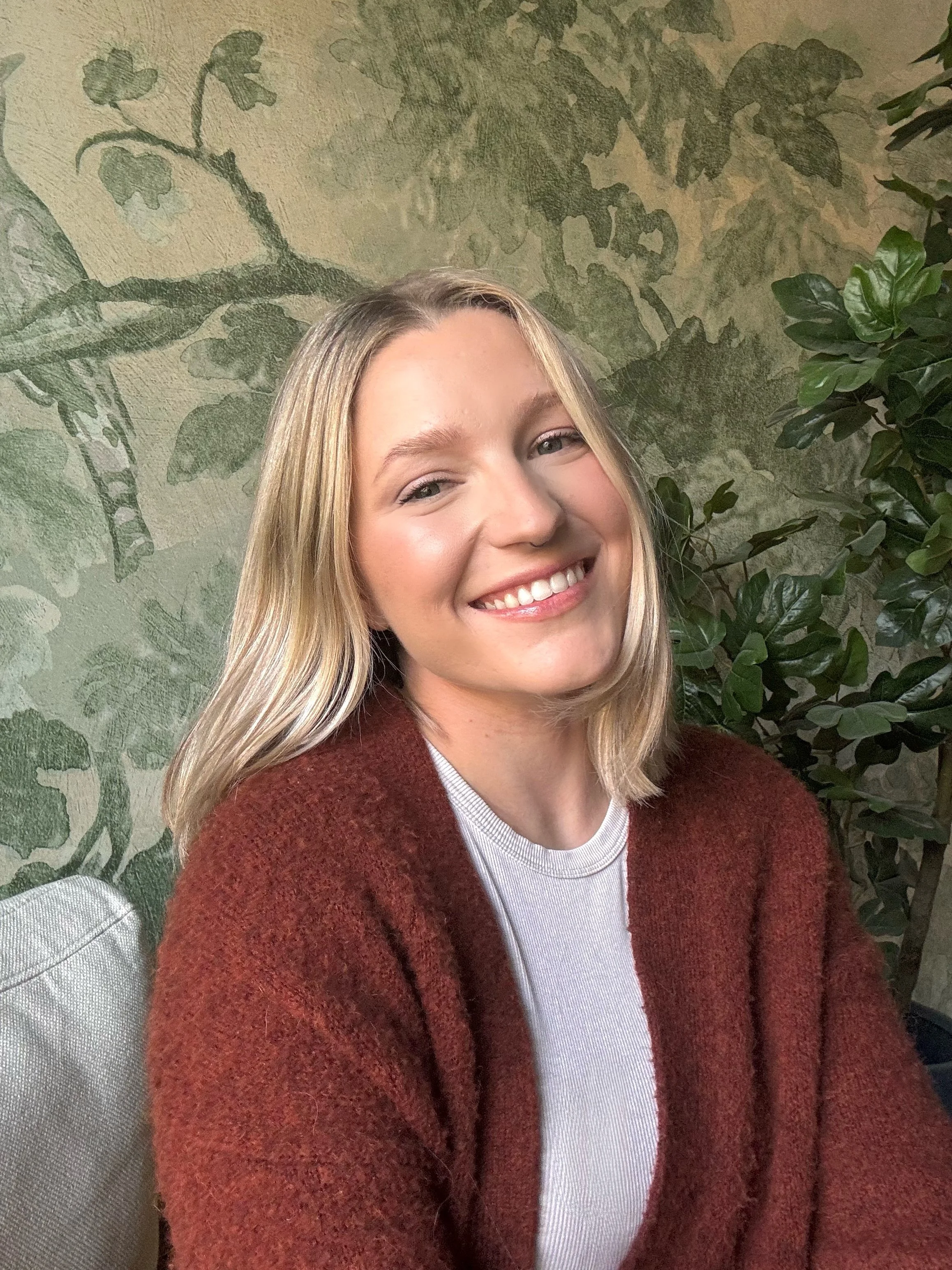 A smiling woman with blonde hair wearing a maroon sweater and a white shirt, sitting next to green leafy plants and a textured wall with a leaf pattern in the background.