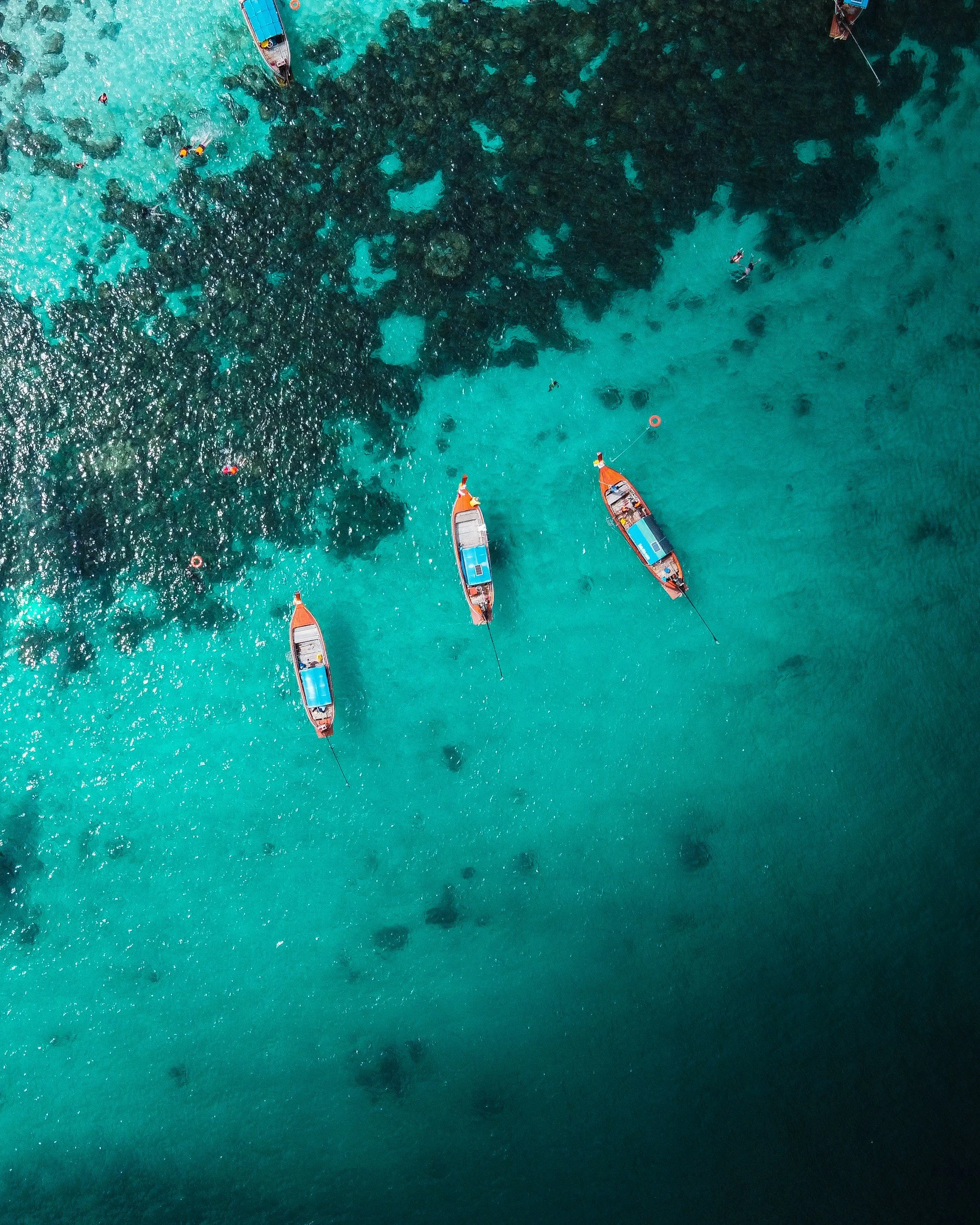Four boats anchored near a coral reef in clear turquoise water, with a few people swimming and snorkeling around the reef.