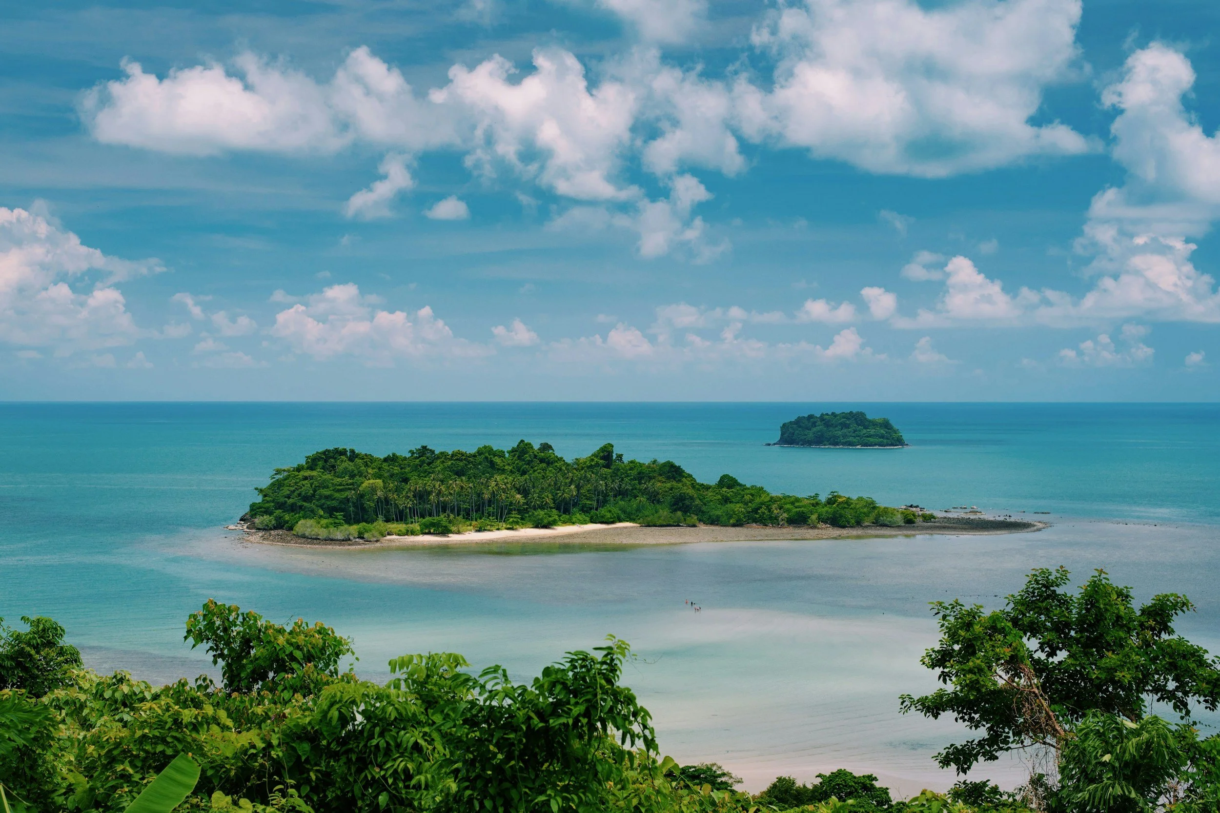 A tropical island surrounded by turquoise waters under a partly cloudy sky, with lush green foliage in the foreground and a smaller island in the background.