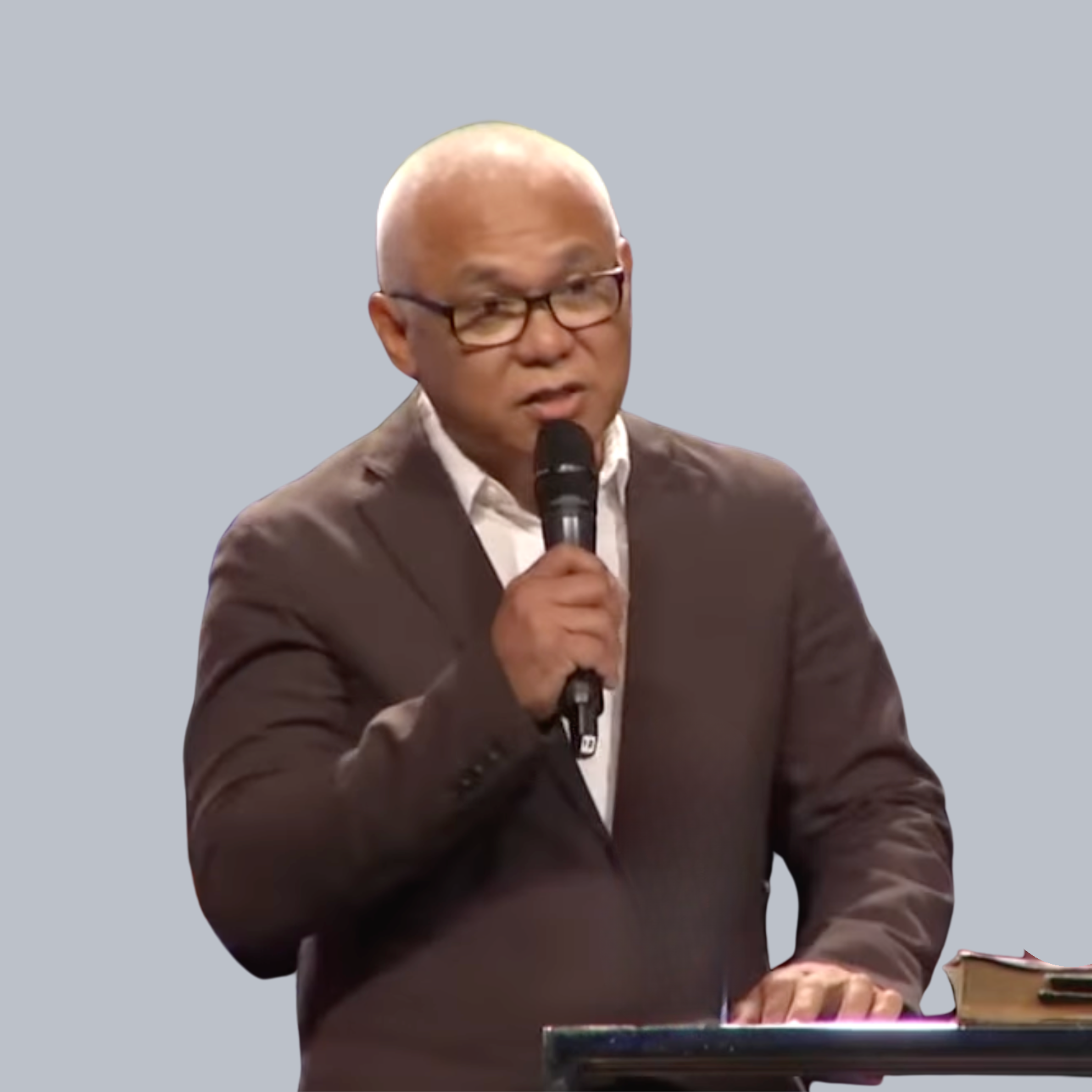 A man in a brown suit and glasses speaking into a microphone.