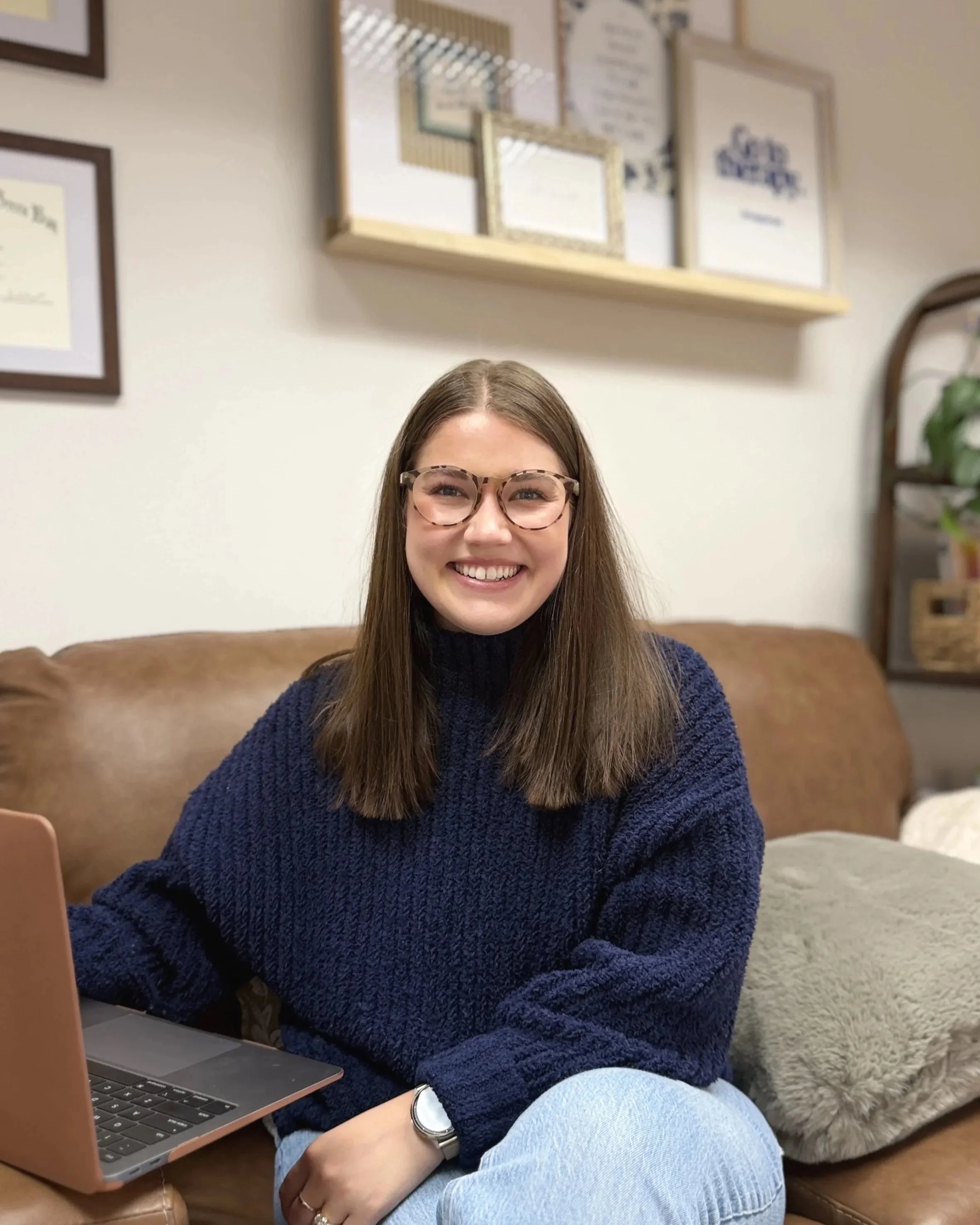 A smiling woman with long brown hair, wearing glasses, a navy blue sweater, and a wristwatch, sitting on a couch in a cozy room with framed certificates and pictures on the wall behind her. There is a laptop in front of her.