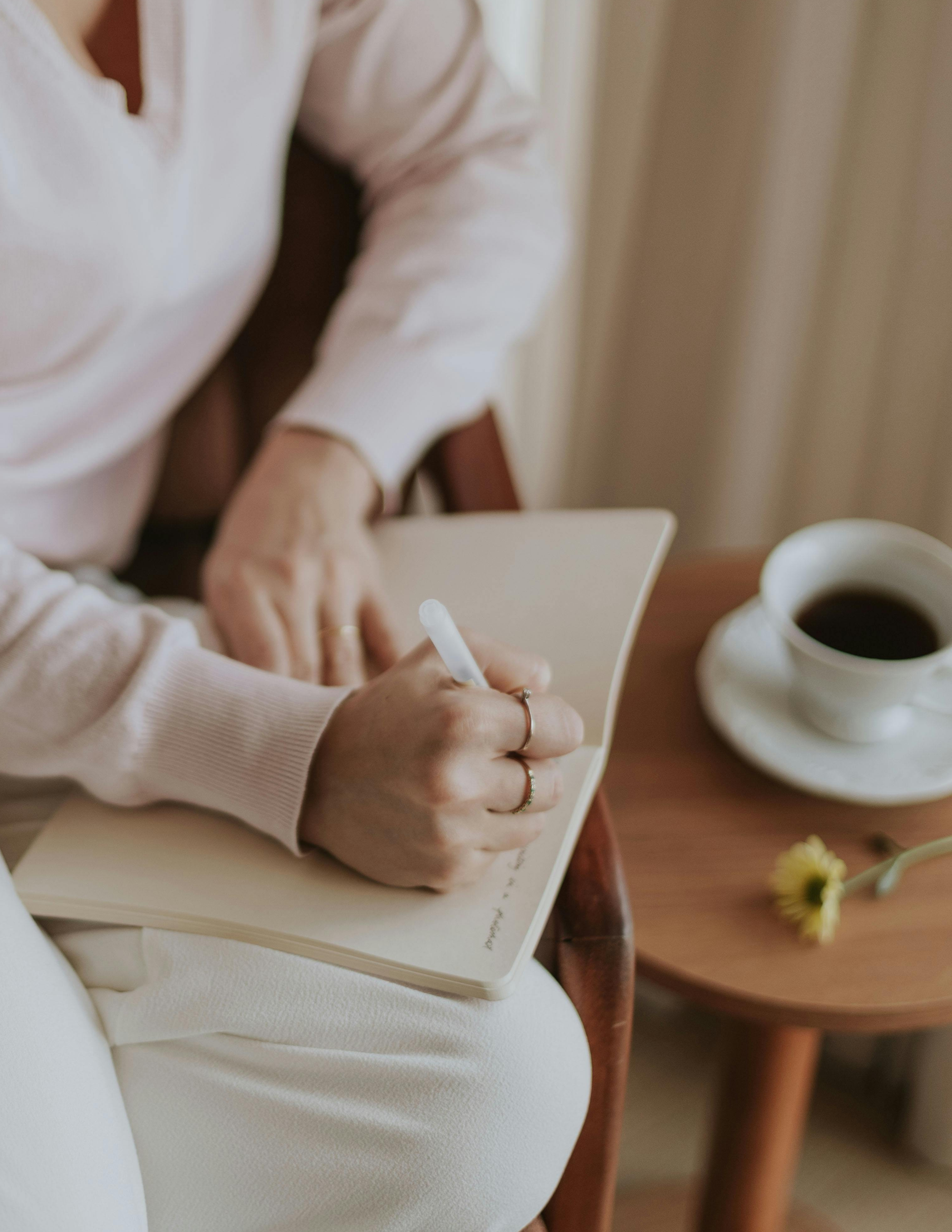 A person writing in a notebook with a pen, seated next to a cup of coffee and a flower on a wooden table.