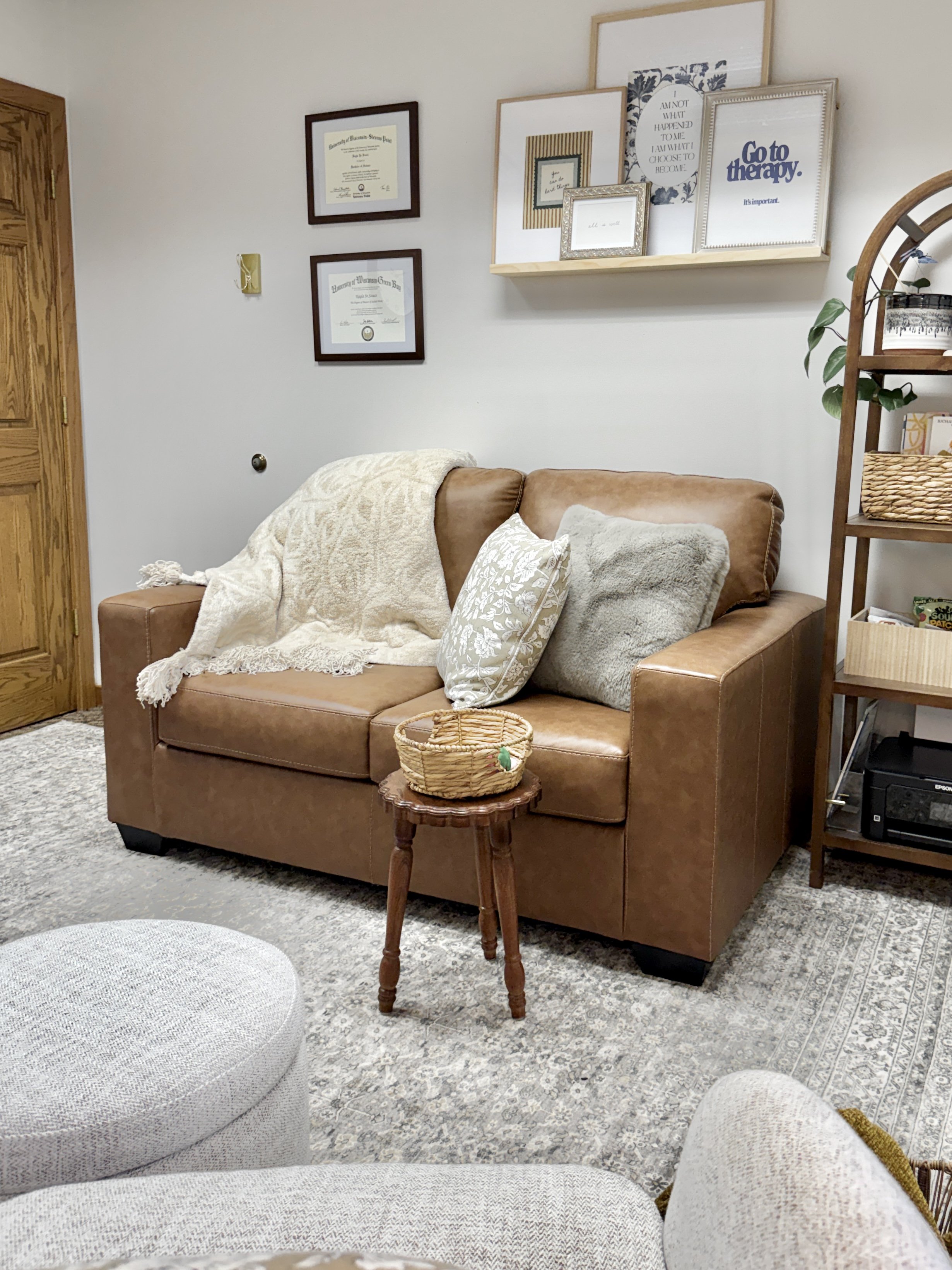 Living room with a leather sofa, patterned pillows, a small wooden side table with a woven basket, and a bookshelf with decorative items. Framed artwork and quotes are on the wall above the sofa and shelf. The floor has a patterned rug.