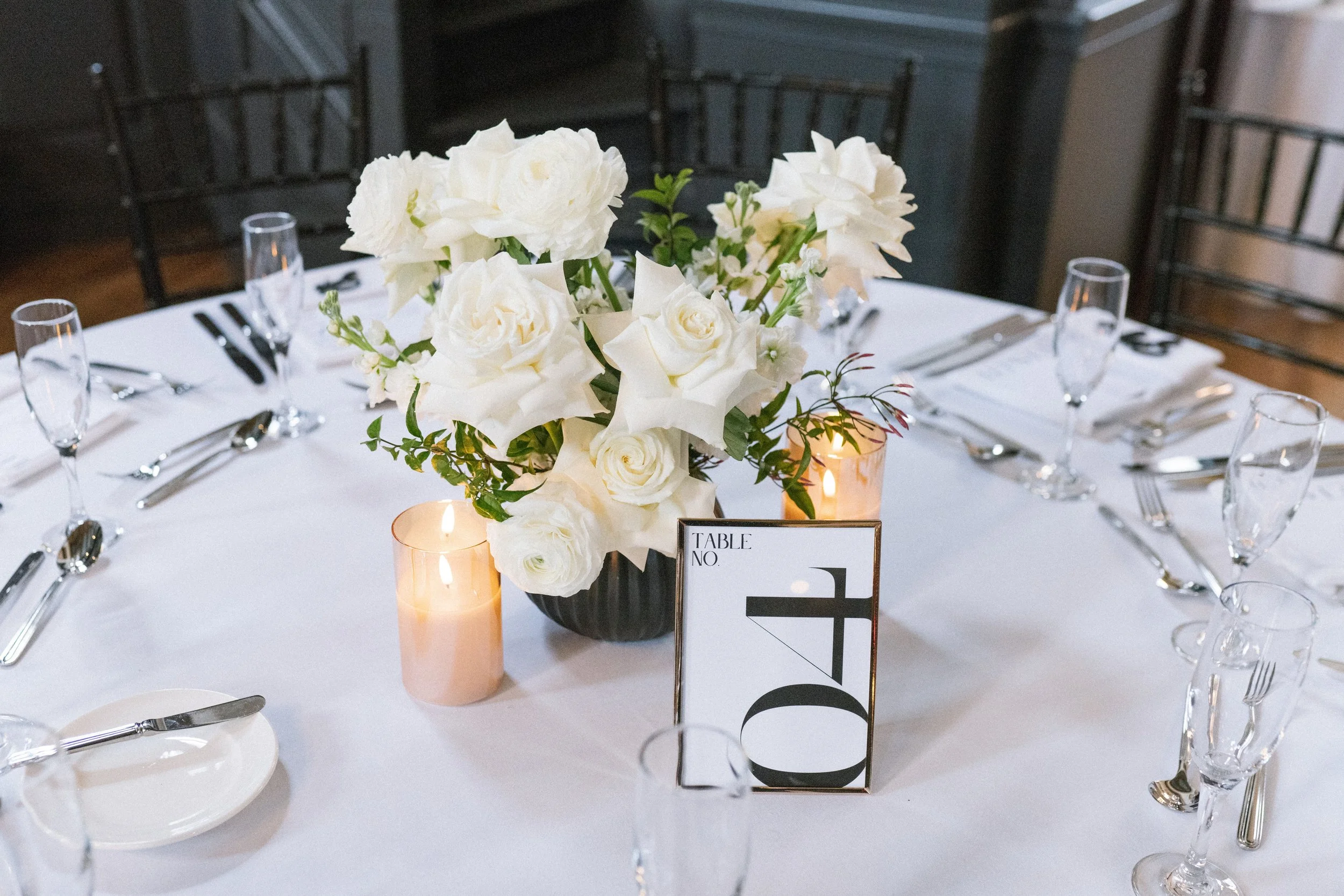 A round banquet table decorated with a large white floral centerpiece, candles, and place settings with glasses, silverware, and white plates