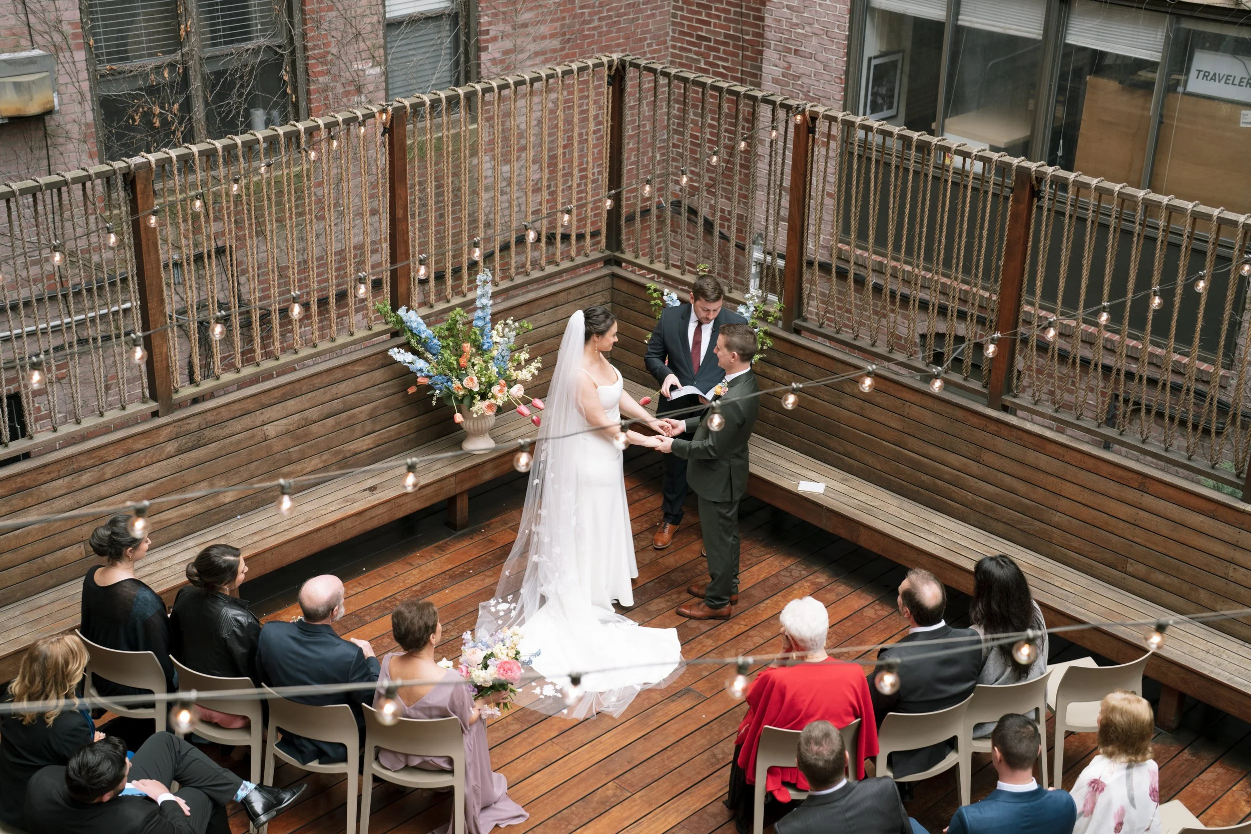 A wedding ceremony taking place on a rooftop patio with wooden flooring, string lights, and brick walls. A bride and groom are holding hands and exchanging vows while a minister officiates. Guests are seated around the couple, and a large floral arrangement is visible on a bench.