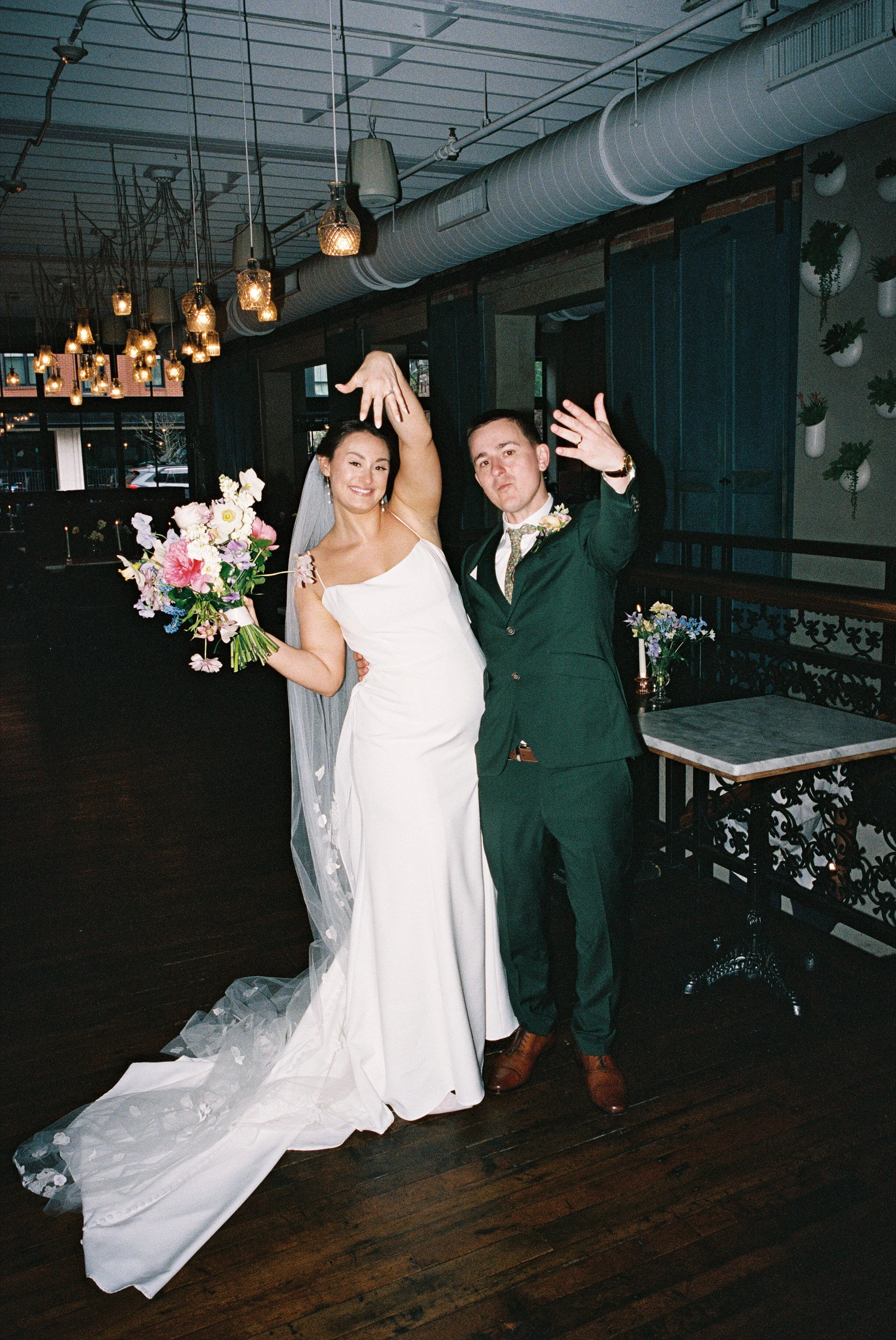 A bride in a white wedding dress holding a colorful bouquet and a groom in a green suit, cheering and celebrating indoors with decorative lighting and plants.