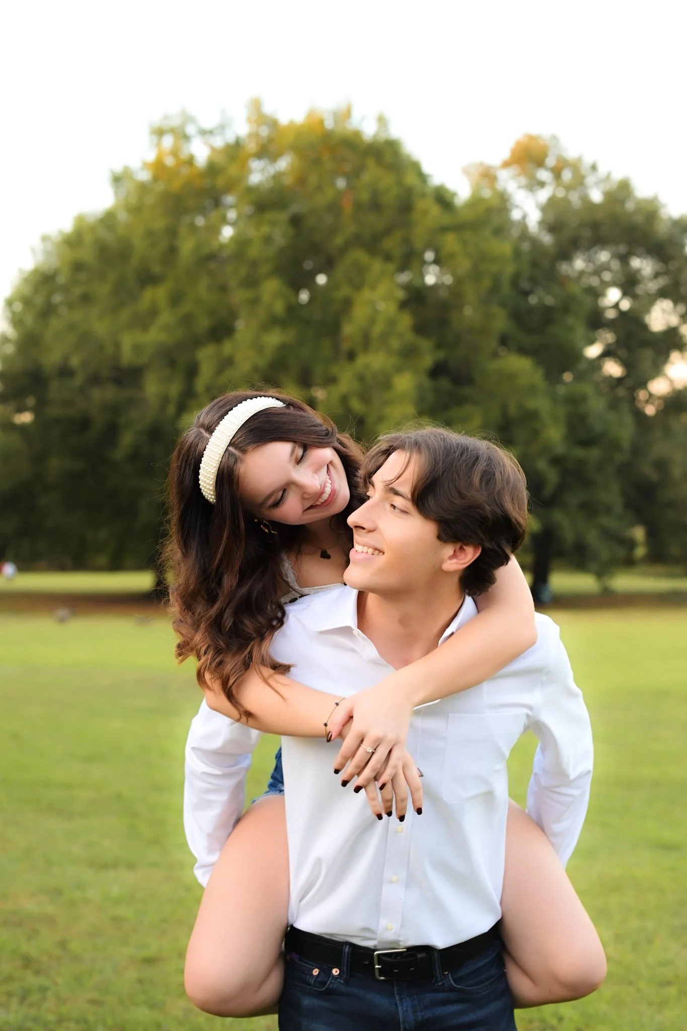 A young couple outdoors in a park, with the woman on the man's back, smiling and looking at each other, surrounded by green trees.
