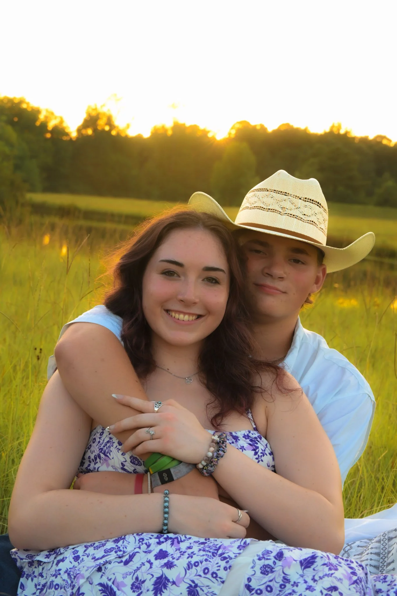 A young couple sitting outdoors in a grassy field during sunset, with the woman smiling and the man wearing a cowboy hat, embracing each other.