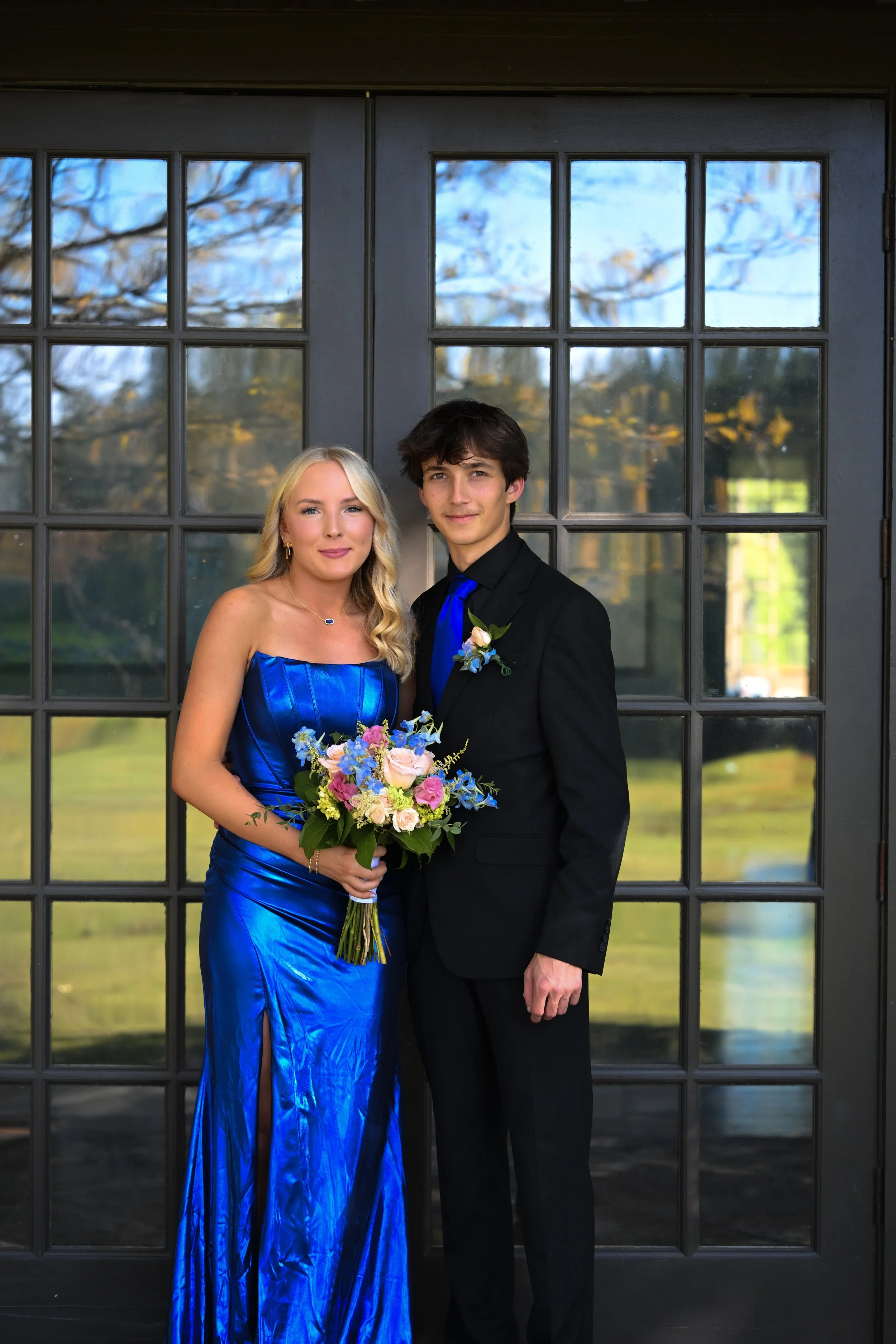 A young woman in a shiny blue gown holding a bouquet of flowers stands next to a young man dressed in a black suit with a blue tie and a boutonnière in front of a black window with a view of trees and a blue sky.