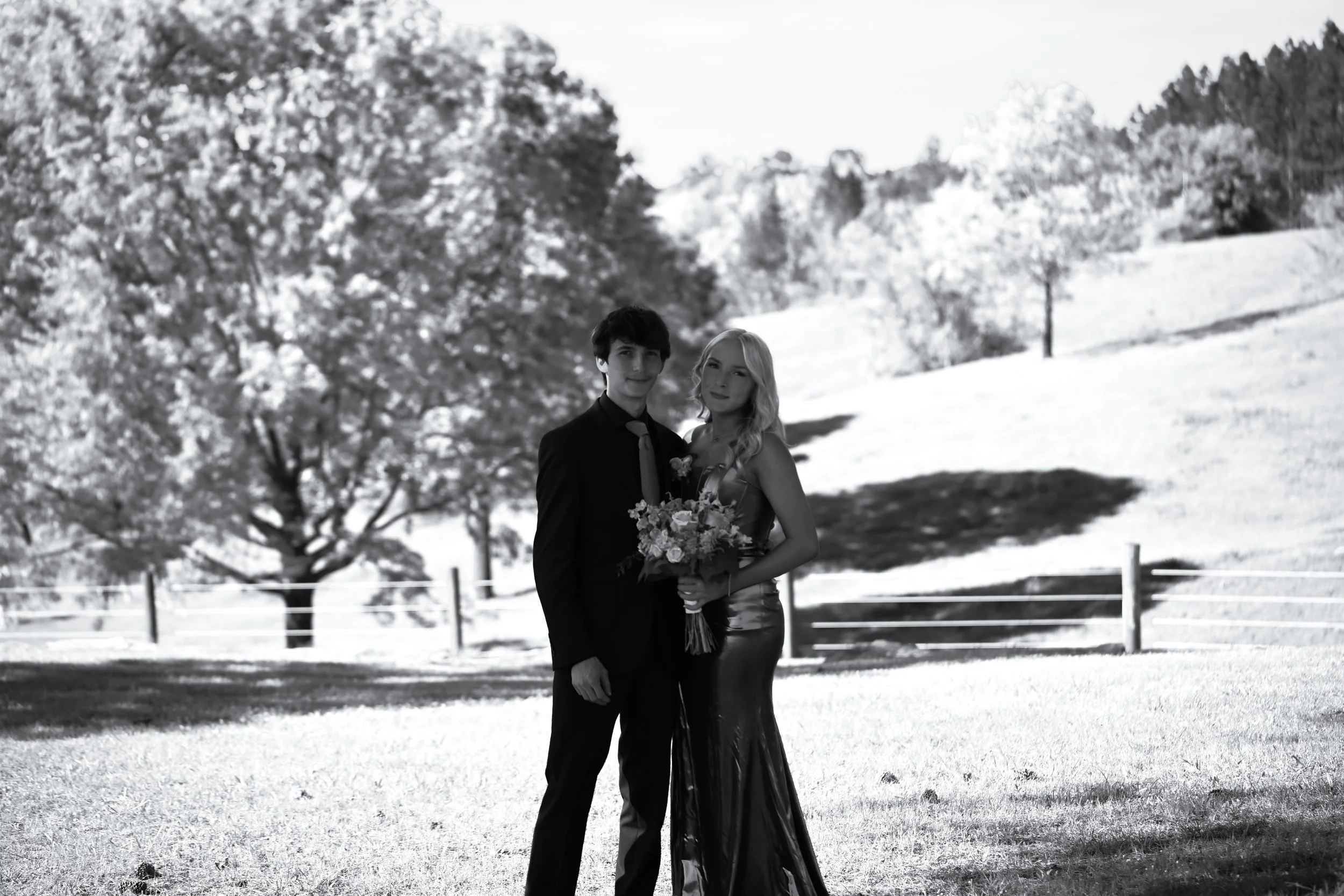 A black and white photo of a young couple dressed for a wedding standing outdoors in a snowy landscape with trees in the background. The woman is holding a bouquet of flowers, and the man is wearing a suit.