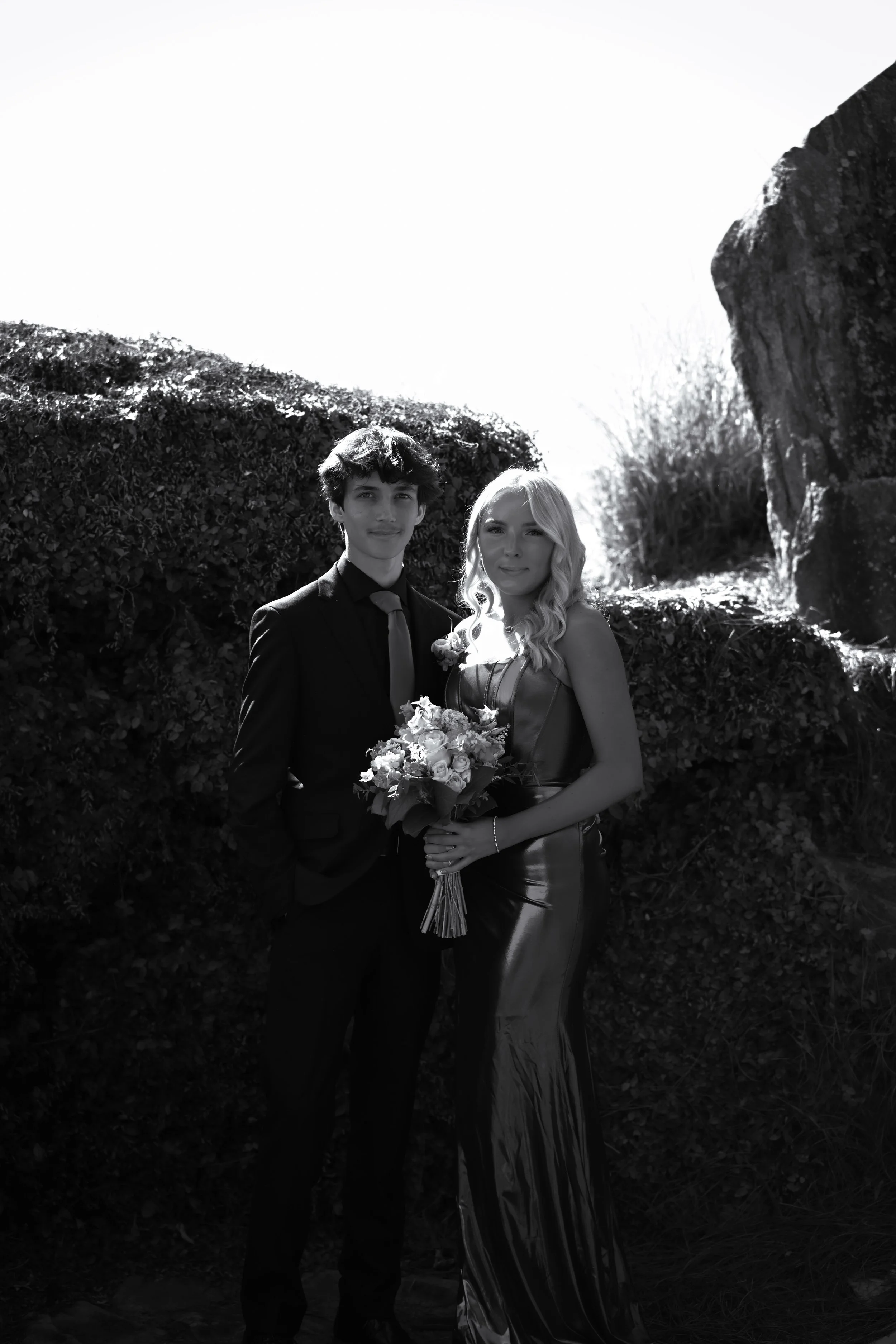 A young man and woman stand outdoors, the woman holding a bouquet of flowers, both dressed in formal attire, with a backdrop of bushes and rocks.