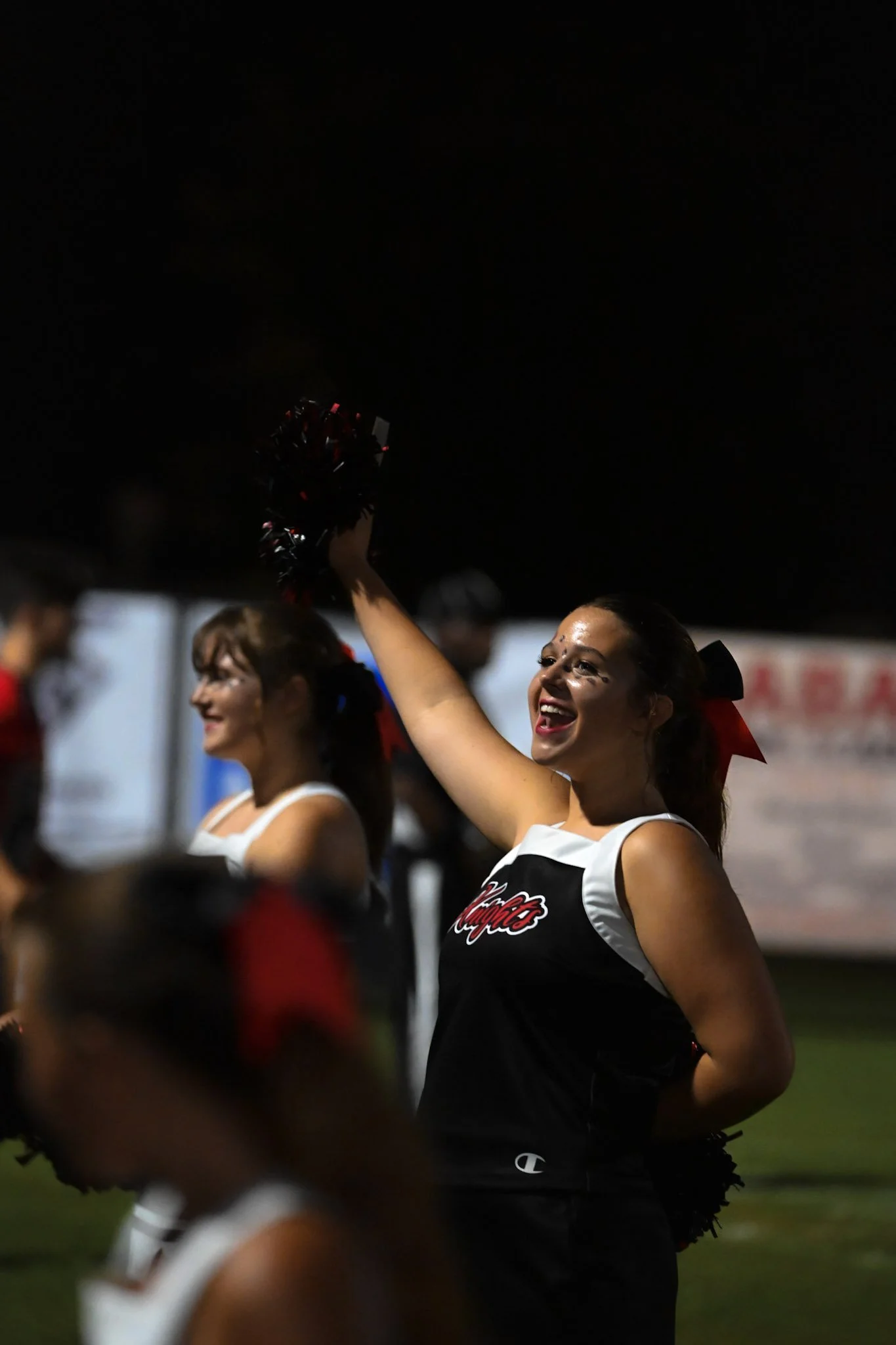 Cheerleaders in uniforms celebrating during a nighttime cheerleading event, smiling and holding pom-poms.