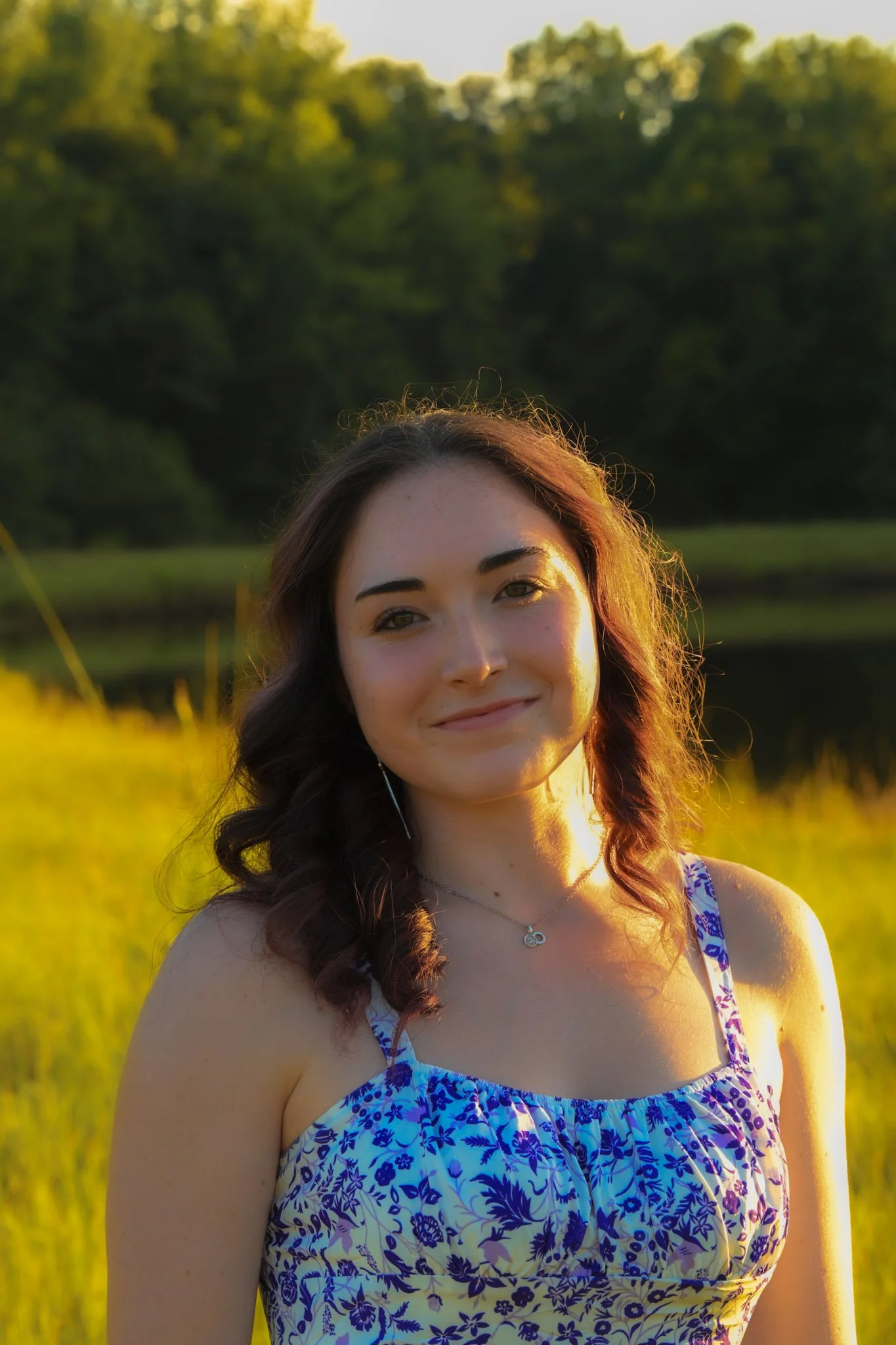 A young woman with brown hair in loose curls, wearing a blue and white floral dress, standing outdoors at sunset near a body of water with trees in the background.