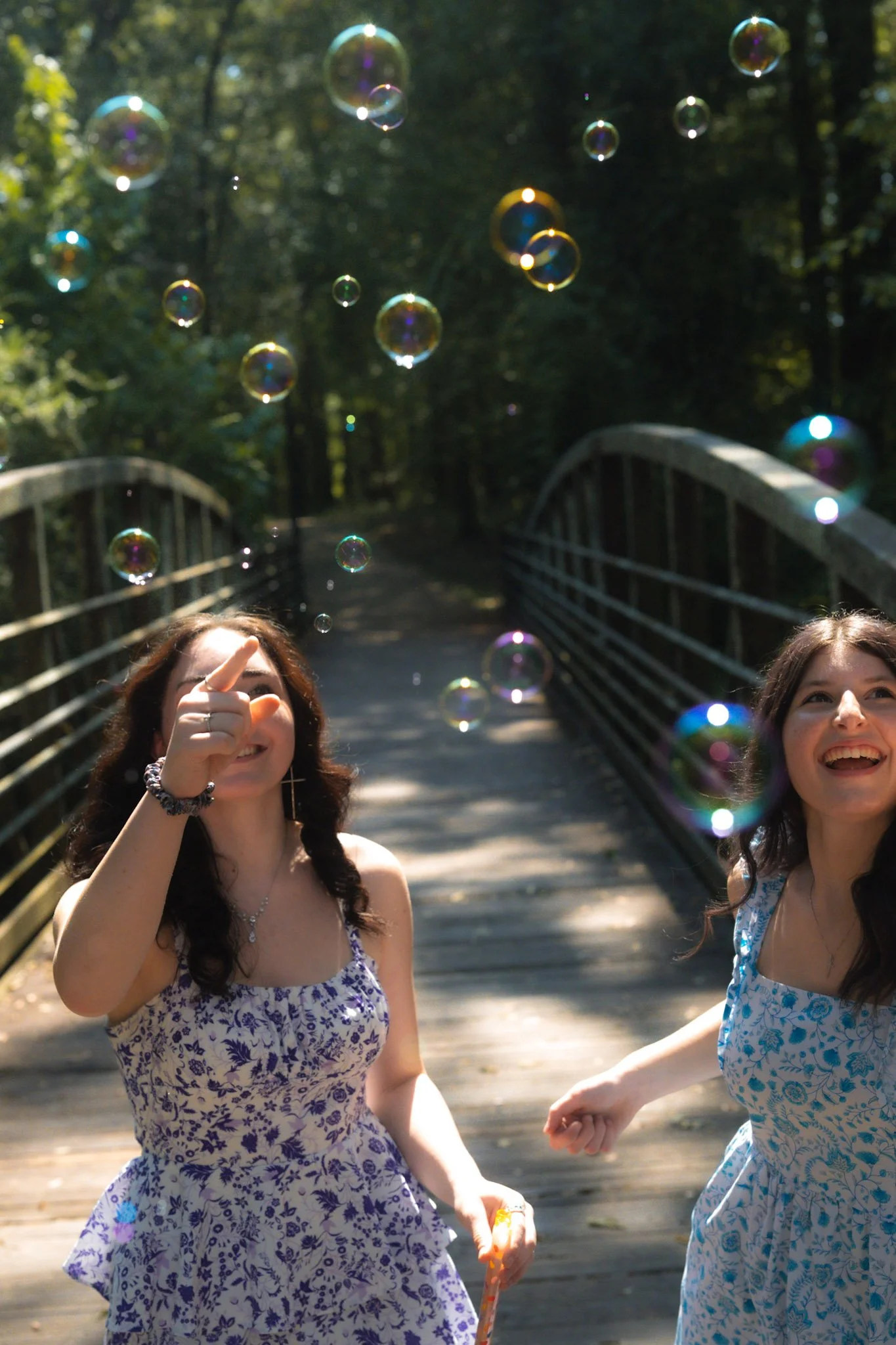 Two young women in dresses smiling and playing with soap bubbles on a wooden bridge in a wooded area.