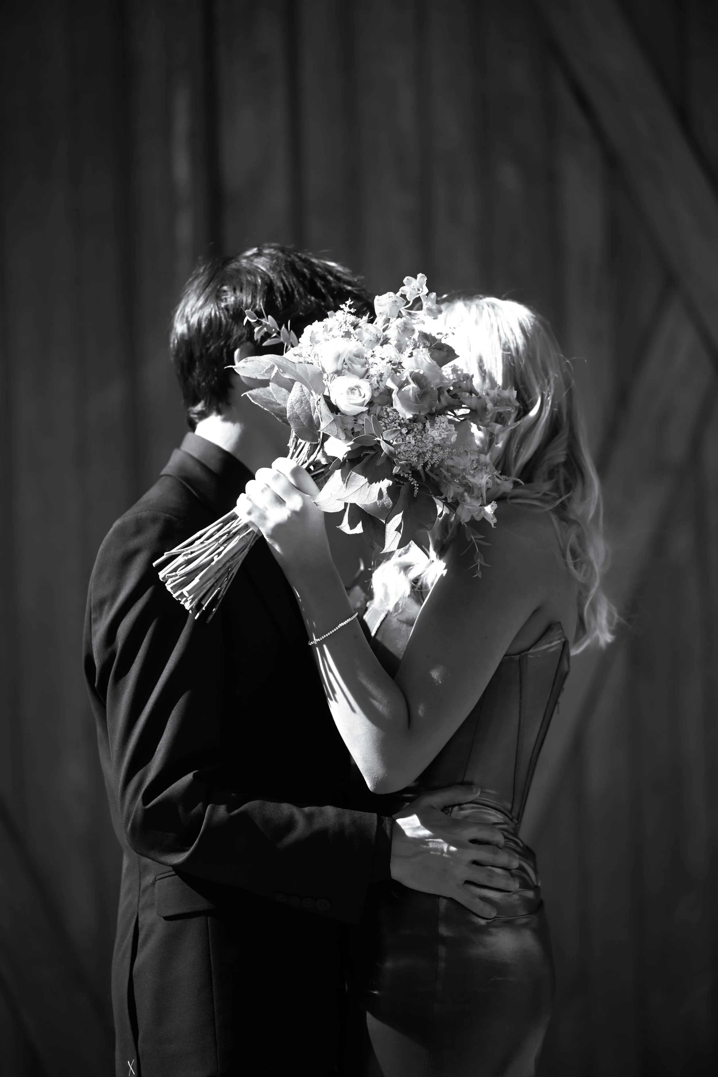A couple dressed in formal attire, with the woman holding a bouquet of flowers, sharing a kiss against a dark background.