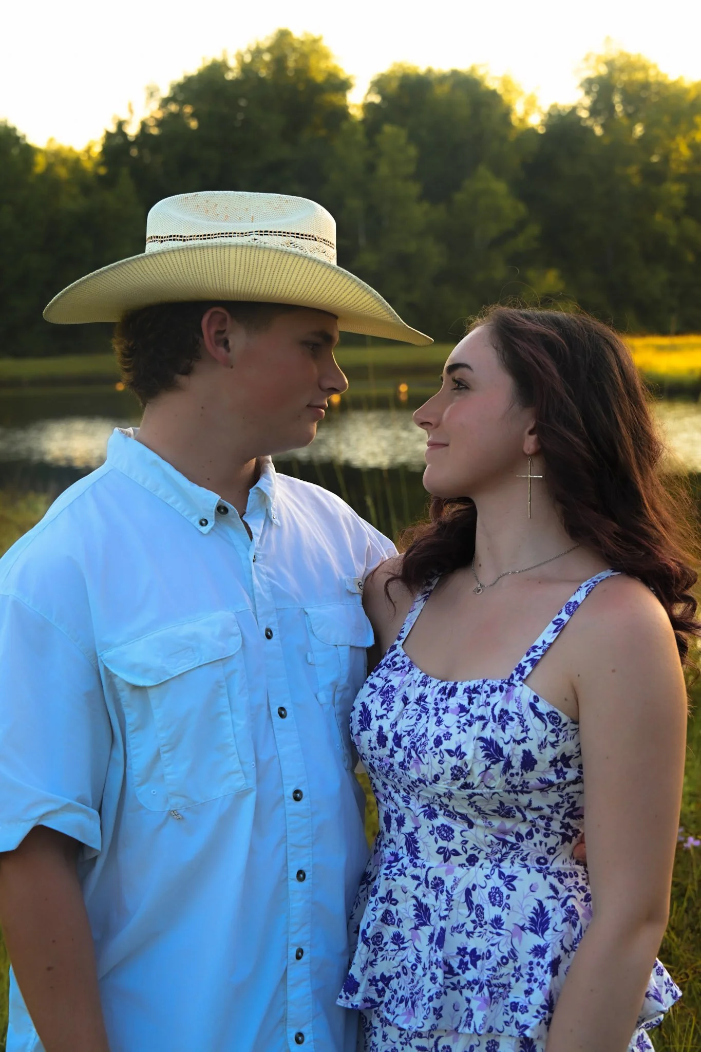 Young couple outdoors near a pond, admiring each other at sunset. The man is wearing a light blue shirt and a wide-brimmed hat, and the woman is in a blue and white floral dress with earrings.