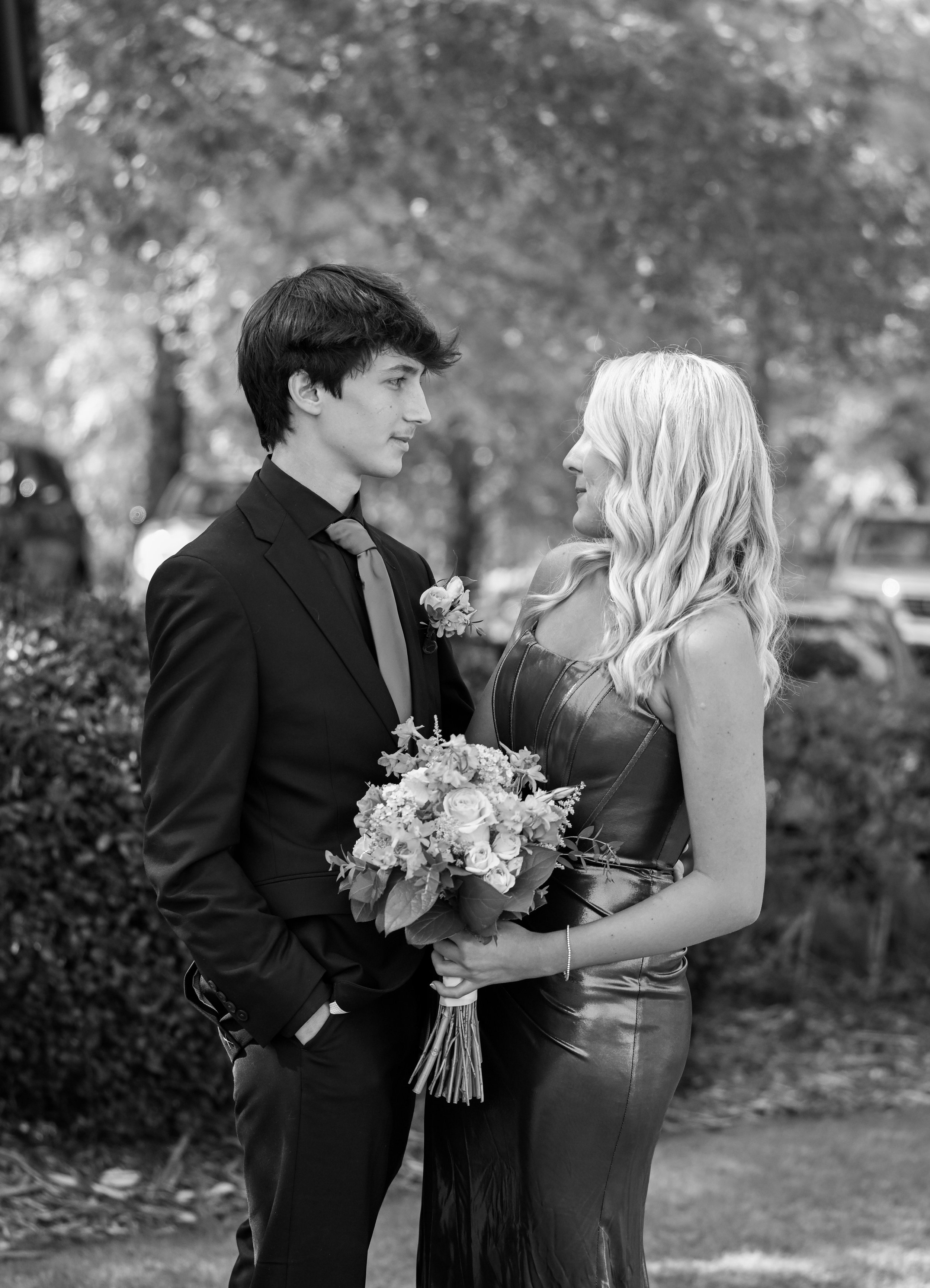 A young man and woman looking at each other outdoors. The man is in a dark suit with a boutonnière, and the woman is in a shiny dress, holding a bouquet of flowers. They are about to kiss.
