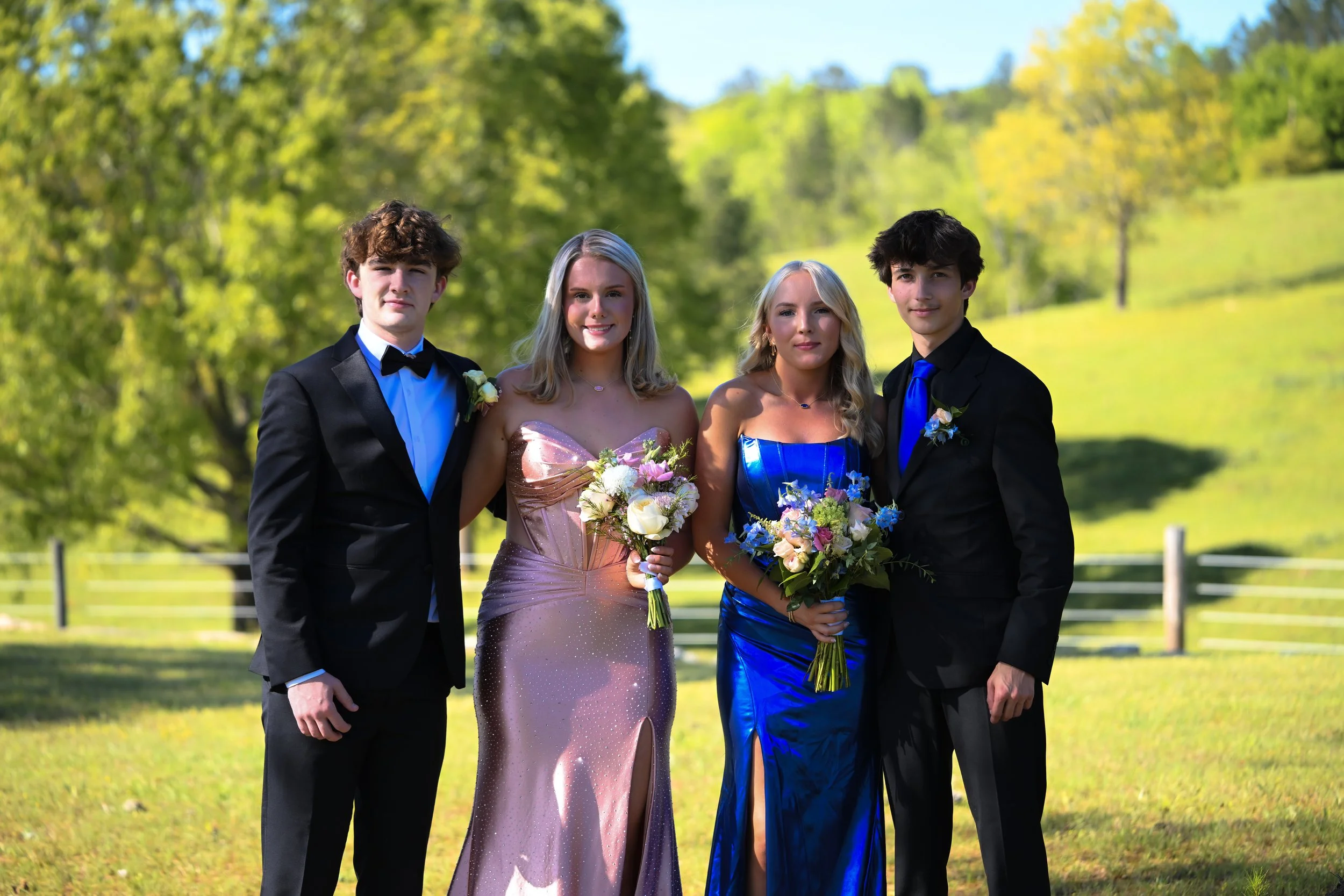 Four teenagers dressed in formal attire standing outdoors with green trees and grass in the background. Two boys are in black tuxedos with blue shirts and ties, and two girls are in elegant dresses holding bouquets of flowers.