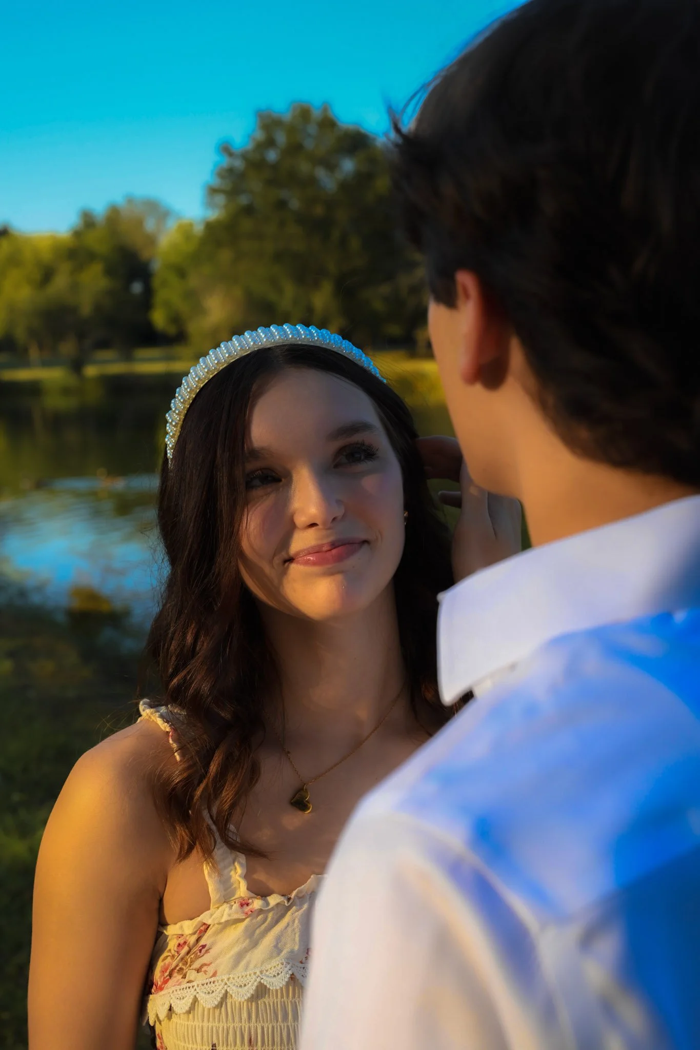 A young woman wearing a pearl headband and a cream dress with lace details looks lovingly at a young man with dark hair, who is facing her and wearing a white shirt, outdoors near a lake surrounded by trees during golden hour.