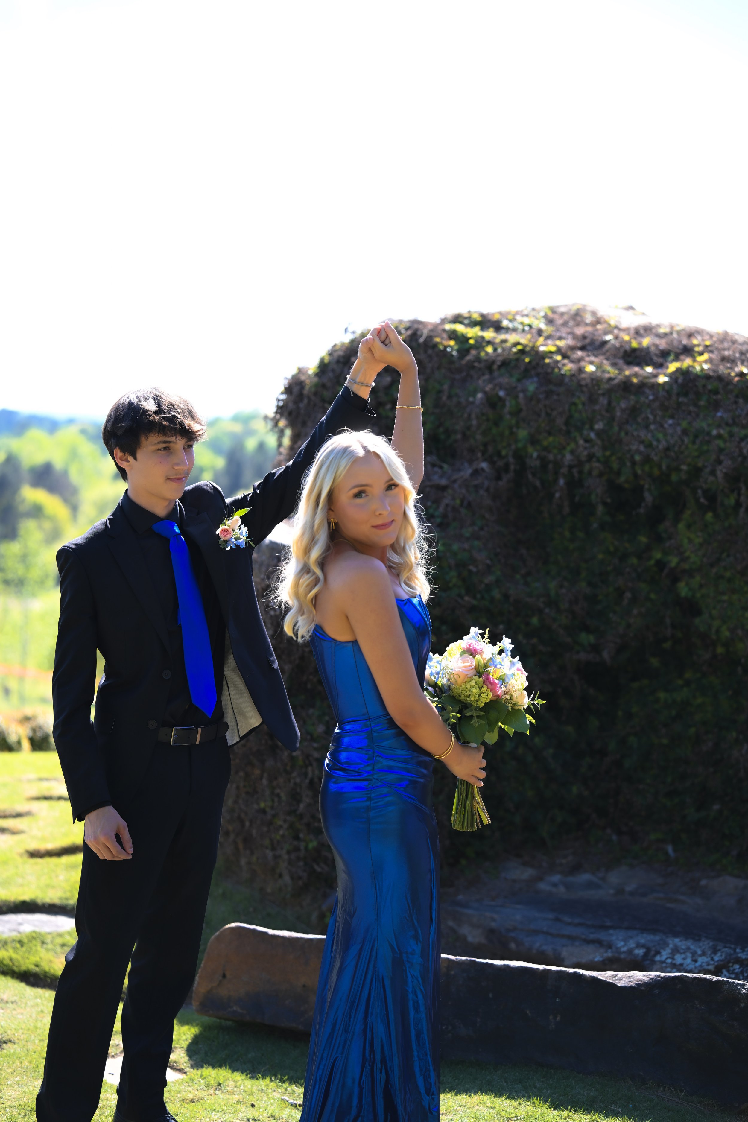 A young man and woman stand outdoors on a sunny day, holding hands with the woman's arm raised. The man wears a black suit with a blue tie and boutonniere, while the woman wears a strapless blue gown and holds a bouquet of flowers.