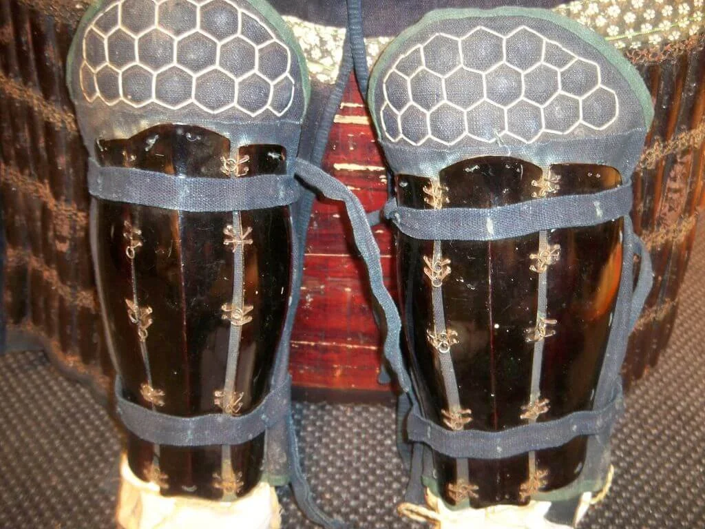 A pair of black shin guards with gold accents, mounted on a mannequin leg for display.