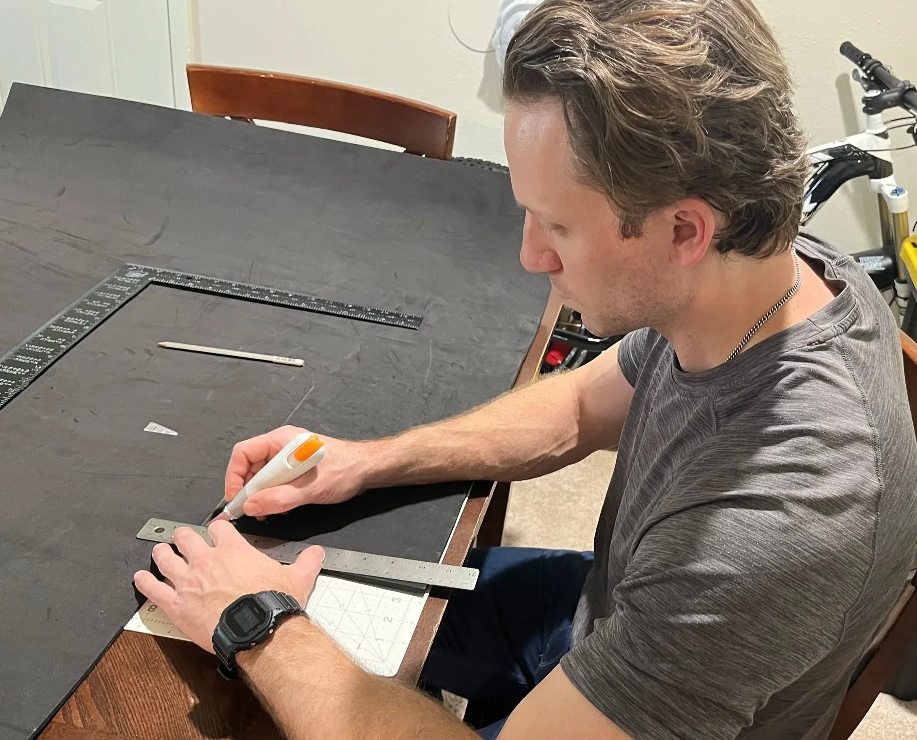 A man with brown hair using an orange and white heat cutter to cut a small piece of metal on a black work surface with a metal ruler and a utility knife, in a room with beige walls and a bicycle in the background.