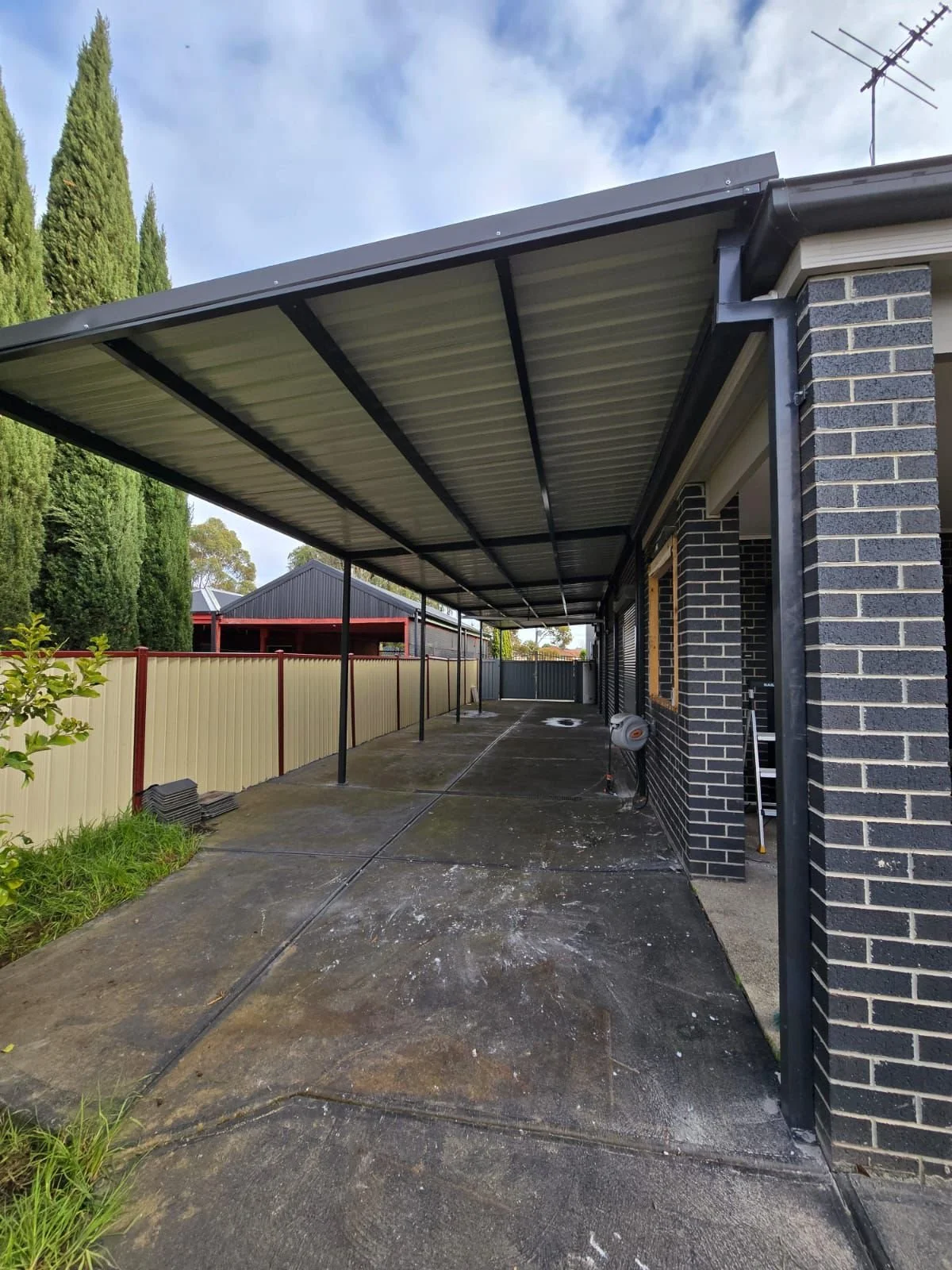 A covered outdoor patio area with a concrete floor, attached to a brick house, some gutters and an antenna on the roof, with a beige fence and tall green trees in the background.