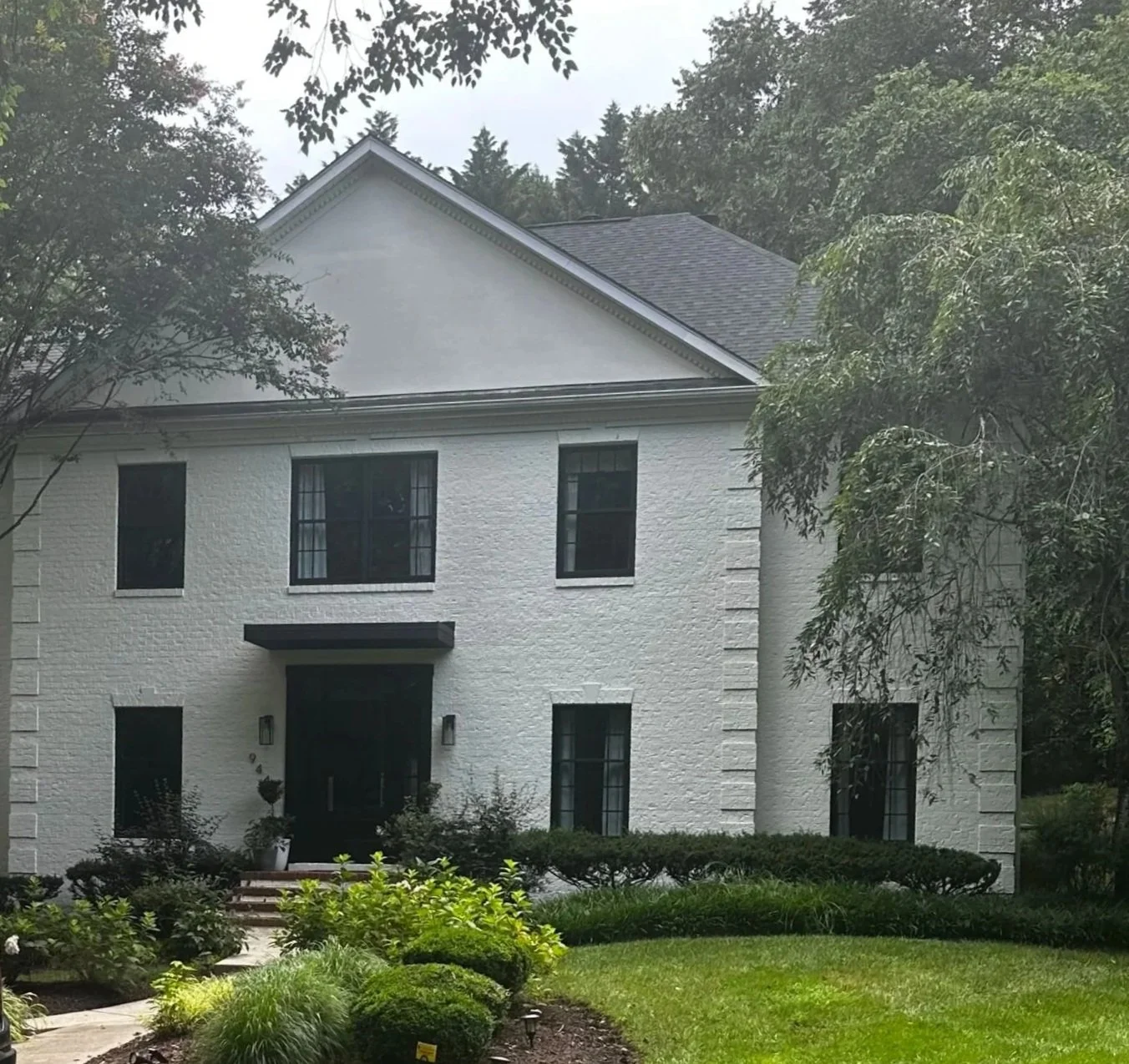 A two-story white brick house with black window frames and a black front door. The house is surrounded by greenery, including trees, shrubs, and a well-maintained lawn.