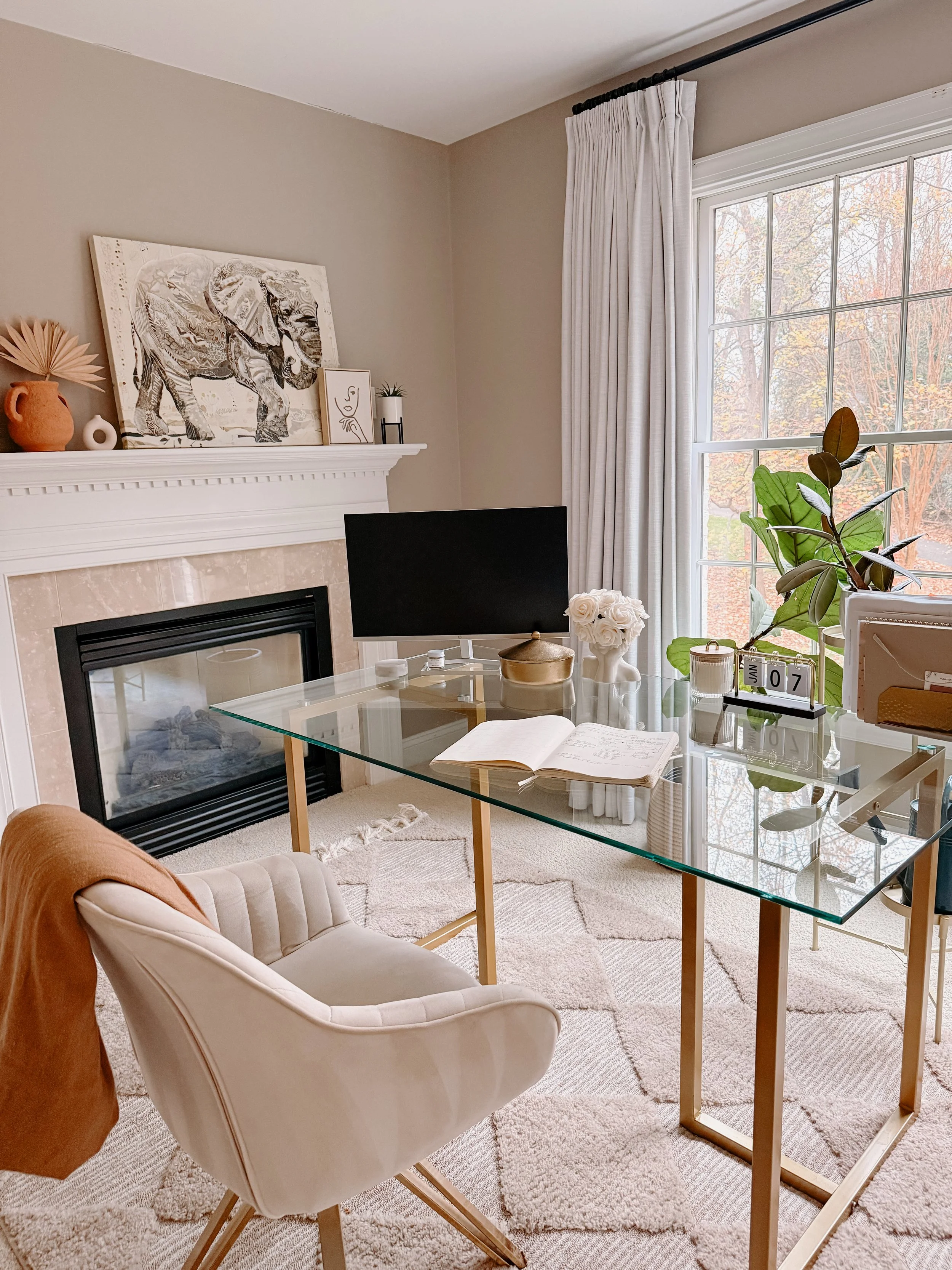 Cozy home office with glass desk, beige armchair, fireplace, window with white curtains, and decorative items including a plant, artwork, and a monitor.