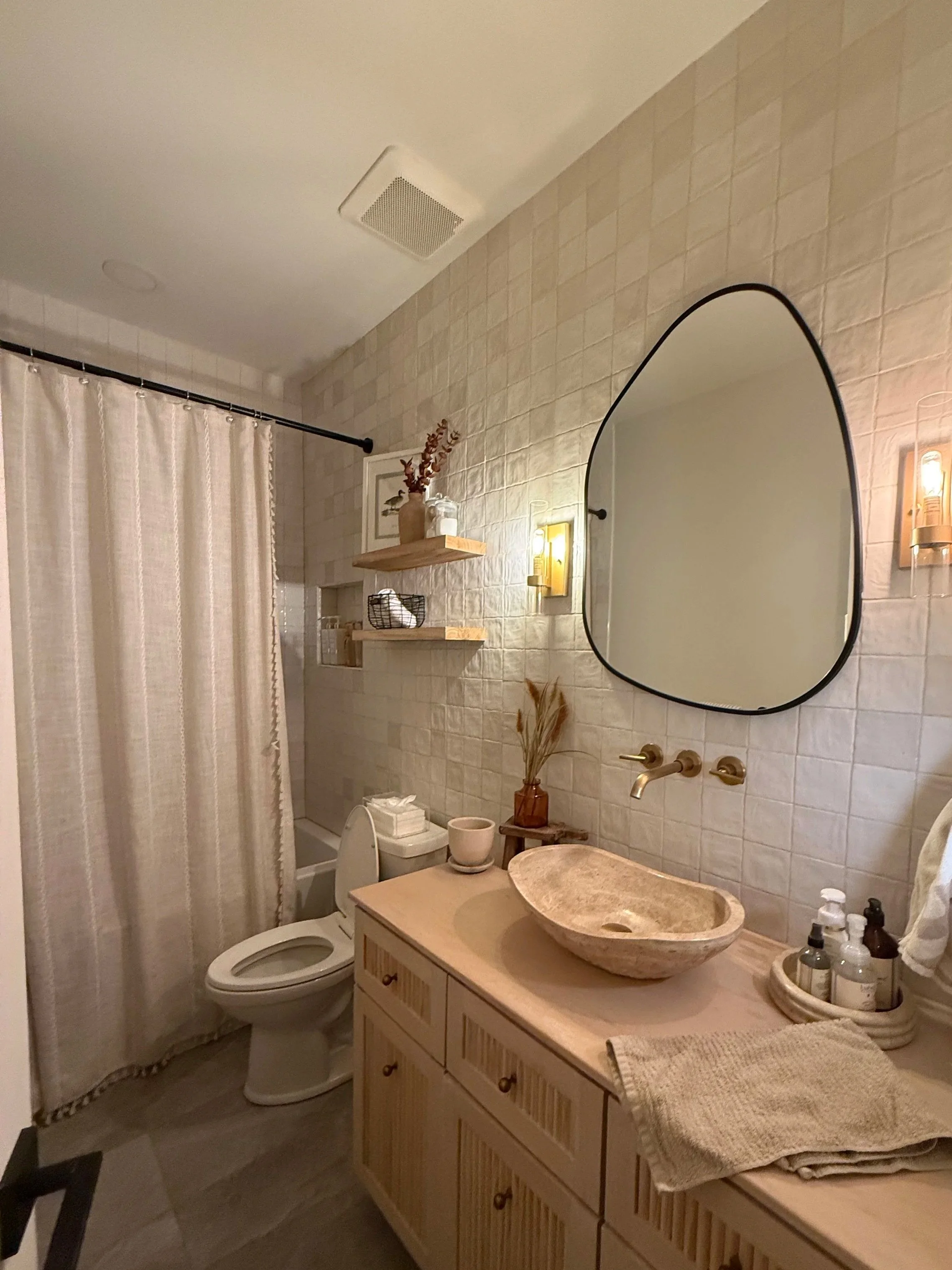 A modern bathroom with beige textured tile walls, a wooden vanity with a stone vessel sink, a round mirror, and a shower area with a curtain. Decor includes shelves with vases, towels, and toiletries.