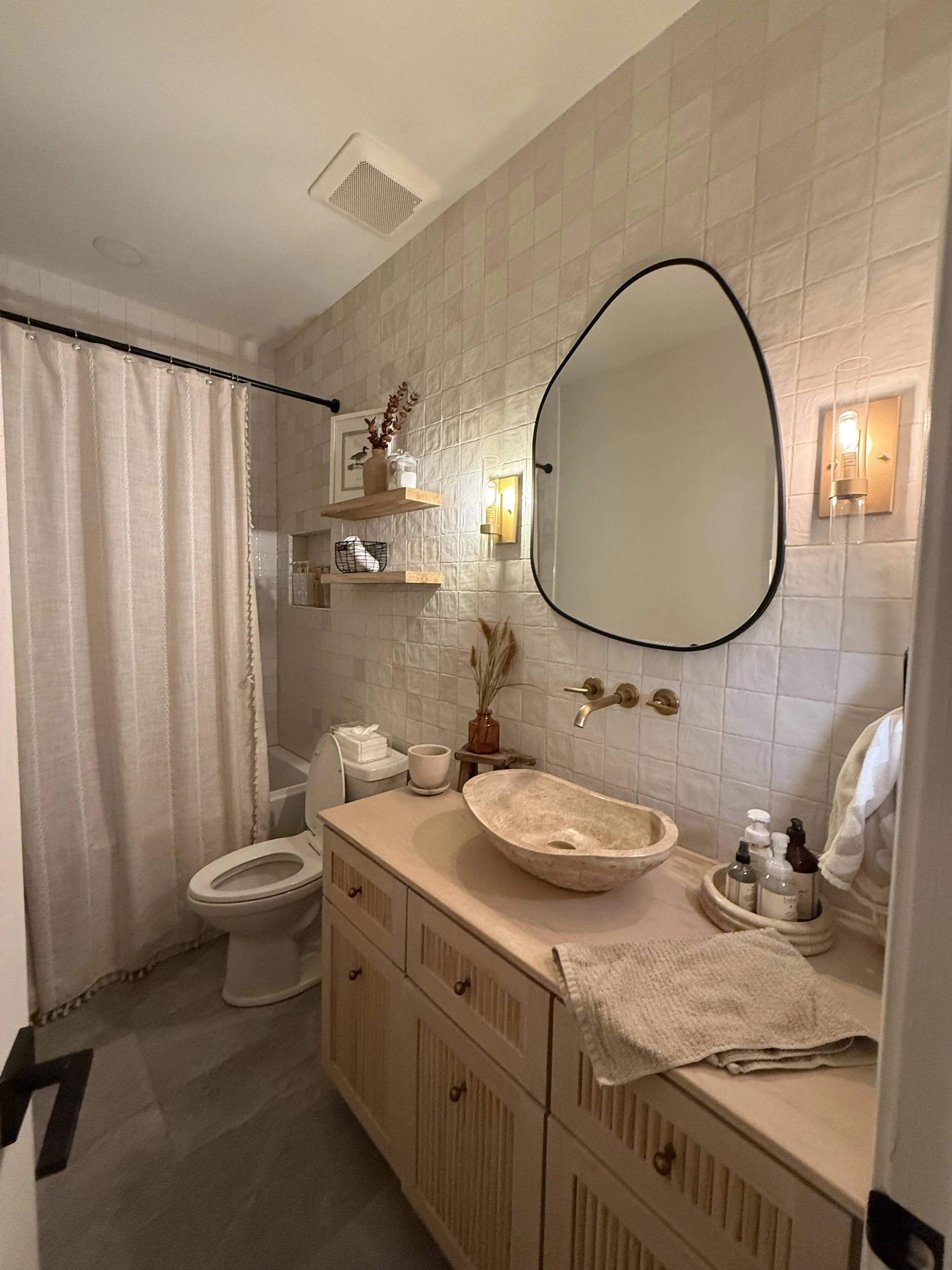 A modern bathroom with a white tiled wall, wooden vanity with a vessel sink, oval mirror, and warm lighting. There's a toilet, a shower curtain, and decorative shelves with plants and accessories.