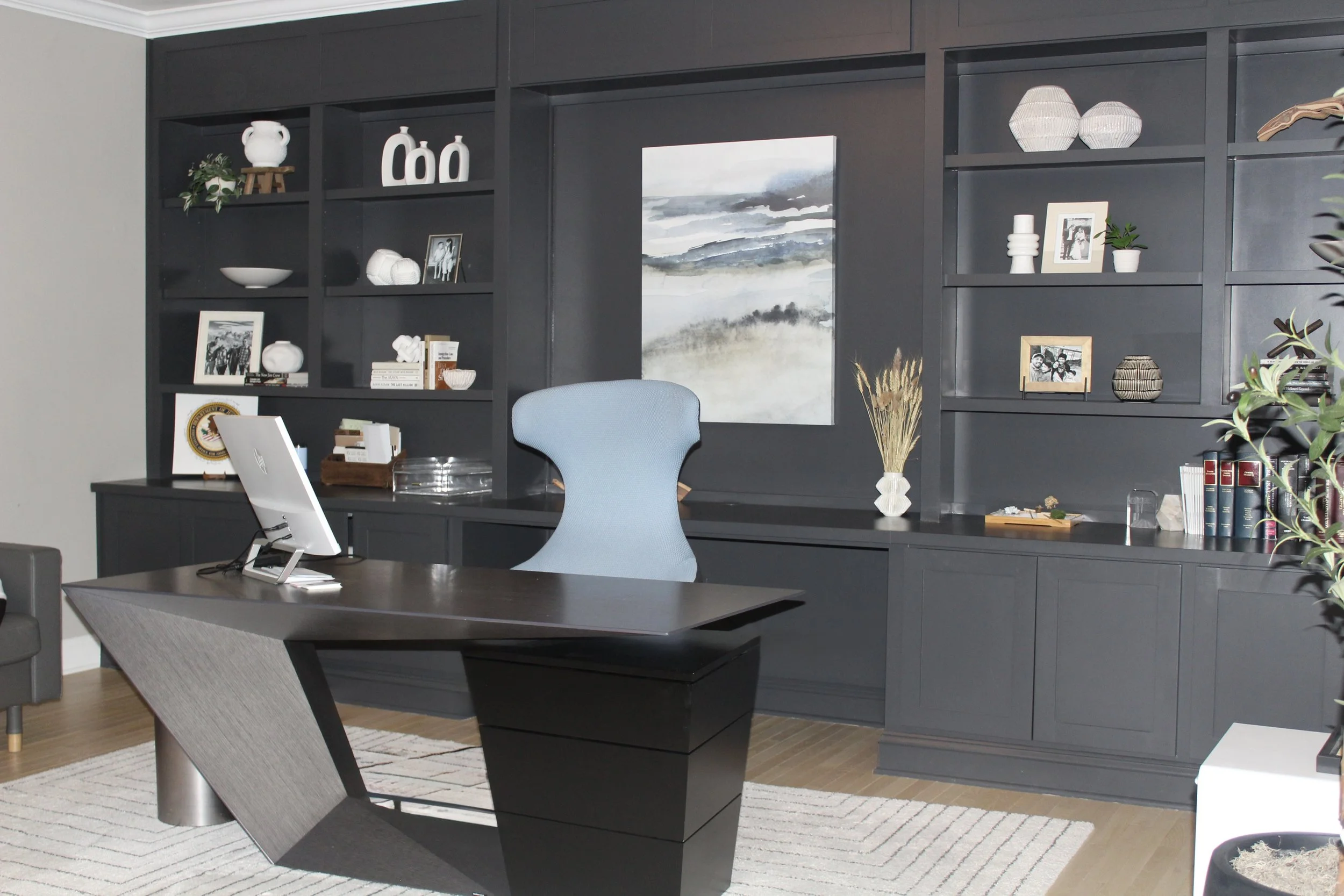 Modern office with a black desk, a light blue chair, a large dark bookshelf with decorative items and framed photos, and a painting on the wall.