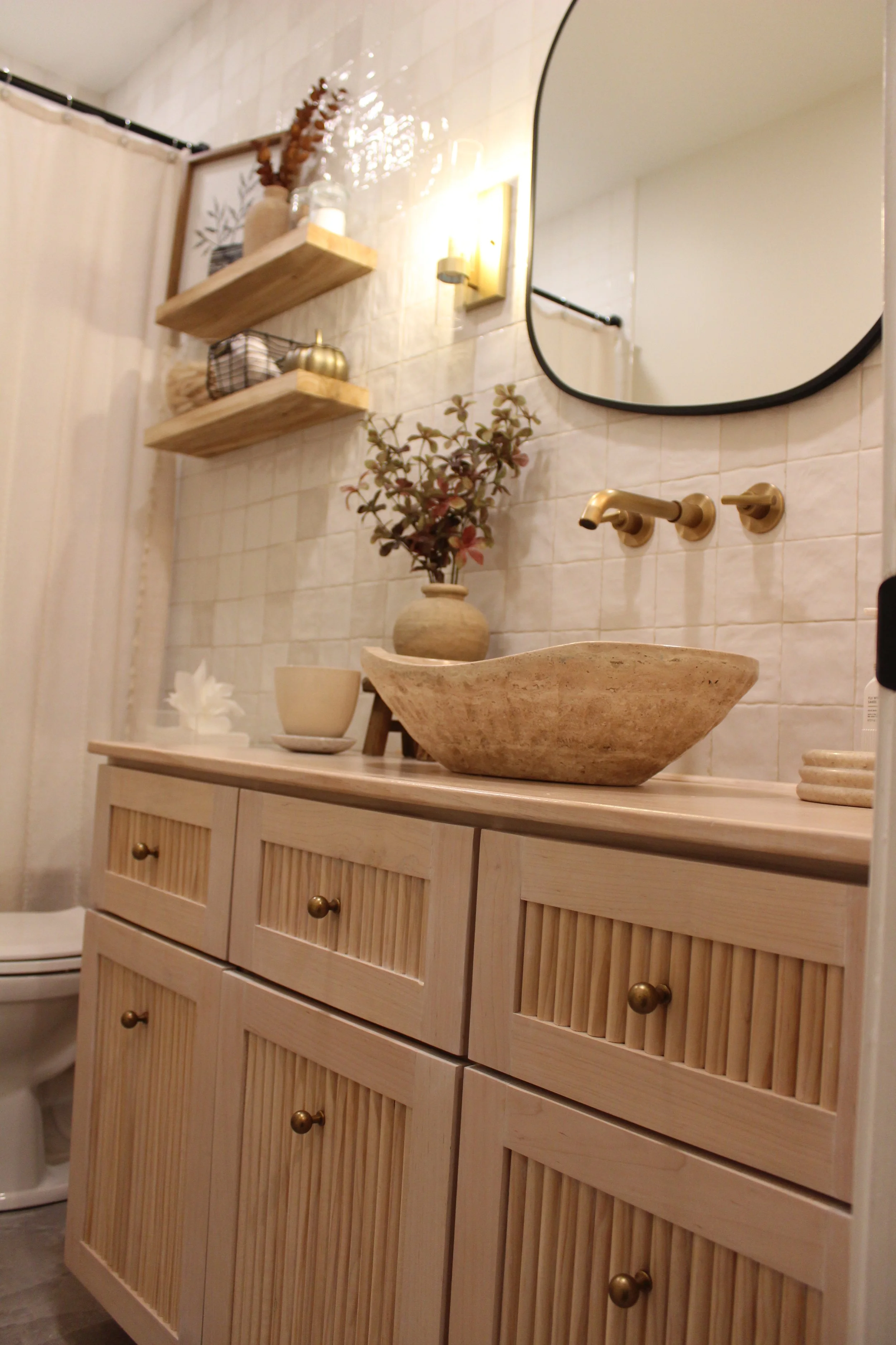 A bathroom vanity with a stone vessel sink, a round mirror, and wooden shelving with decorative items. The wall has beige tiles, and there is a vase with plant, candles, and small decorative objects on the shelves.