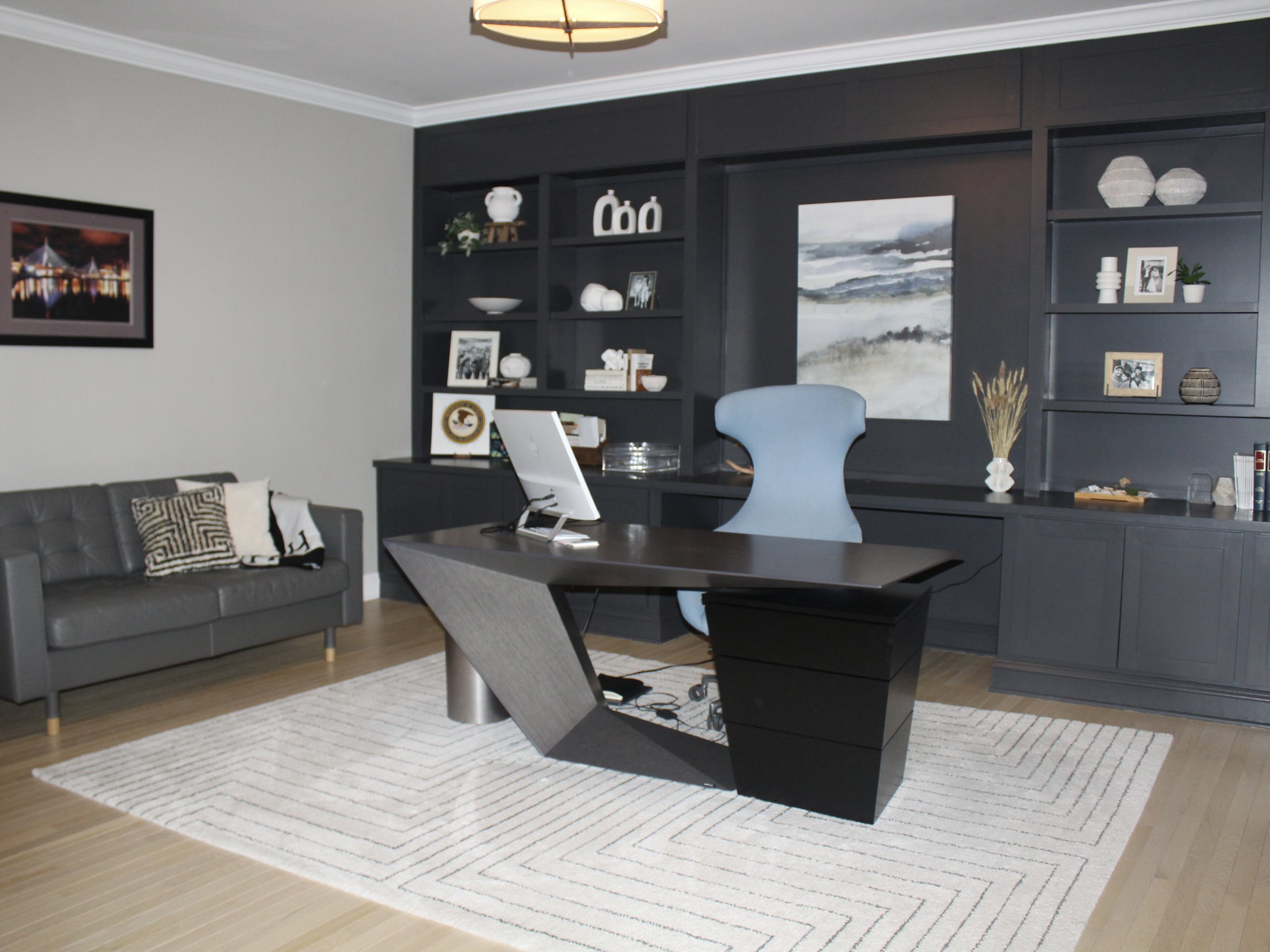 A modern office with a black desk and a blue ergonomic chair, a gray sofa with patterned pillows, dark built-in bookshelves with decorative items, framed artwork, and a white rug on light wooden flooring.