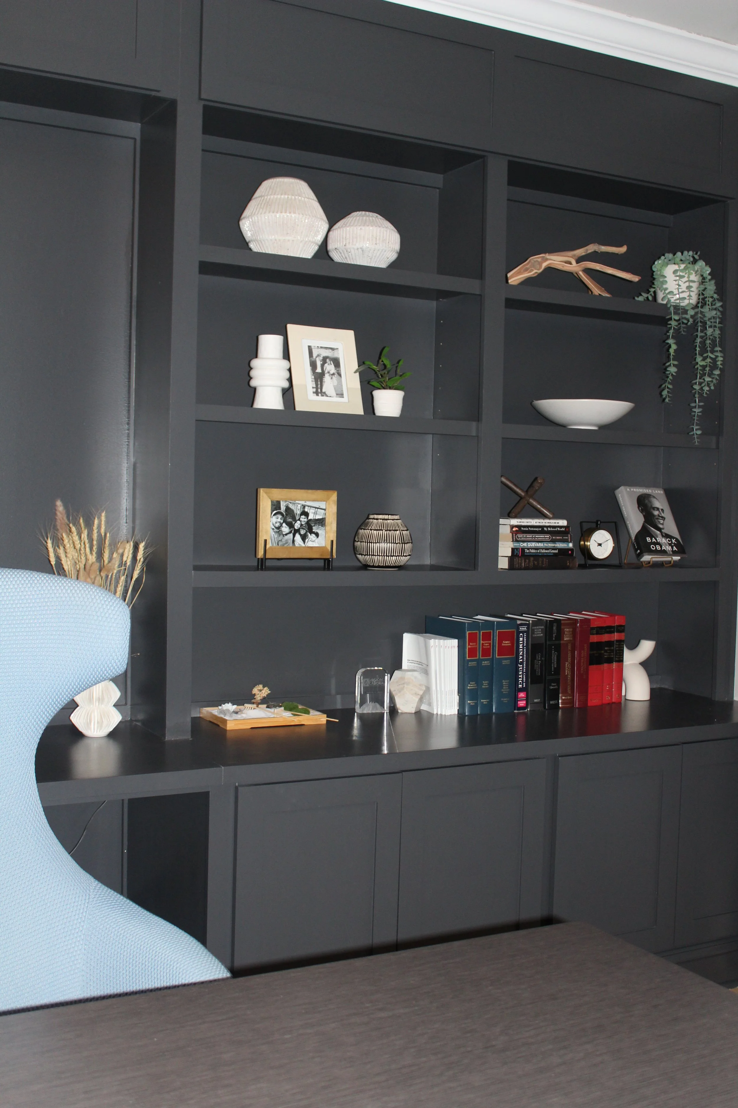 A gray built-in bookshelf with decorative items, books, and plants, next to a blue office chair and a dark wood table.