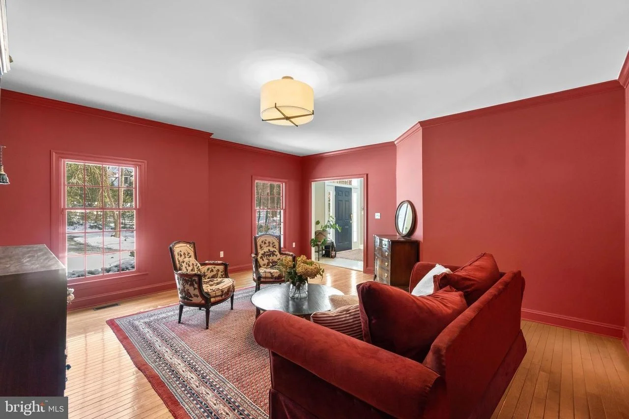 Living room with red walls, hardwood floors, a red velvet sofa, two upholstered armchairs, a round coffee table with flowers, and a window showing snow outside.