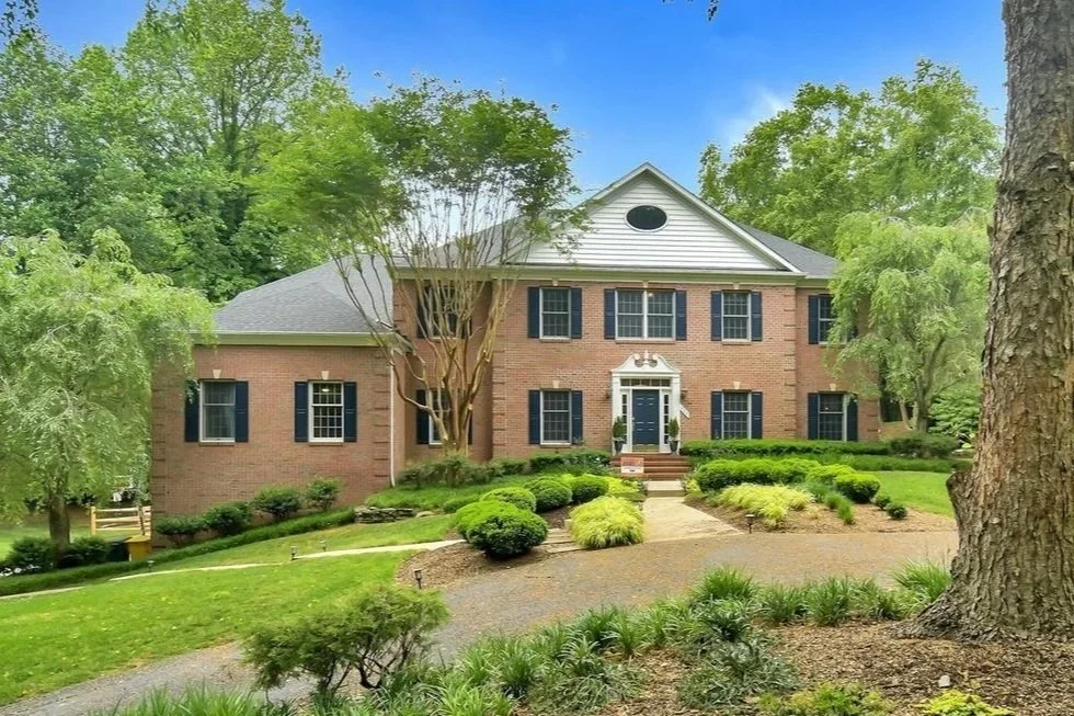 A large brick house with a white front door surrounded by neatly trimmed bushes and trees, set in a lush green landscaped yard under a blue sky.